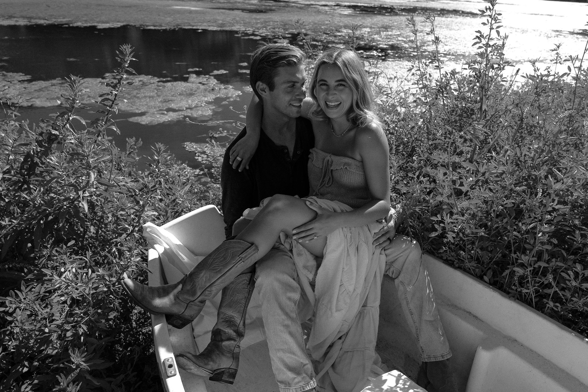 A black and white photo of a smiling couple sitting in a boat surrounded by foliage, with the man wearing casual attire and the woman wearing a dress and cowboy boots.