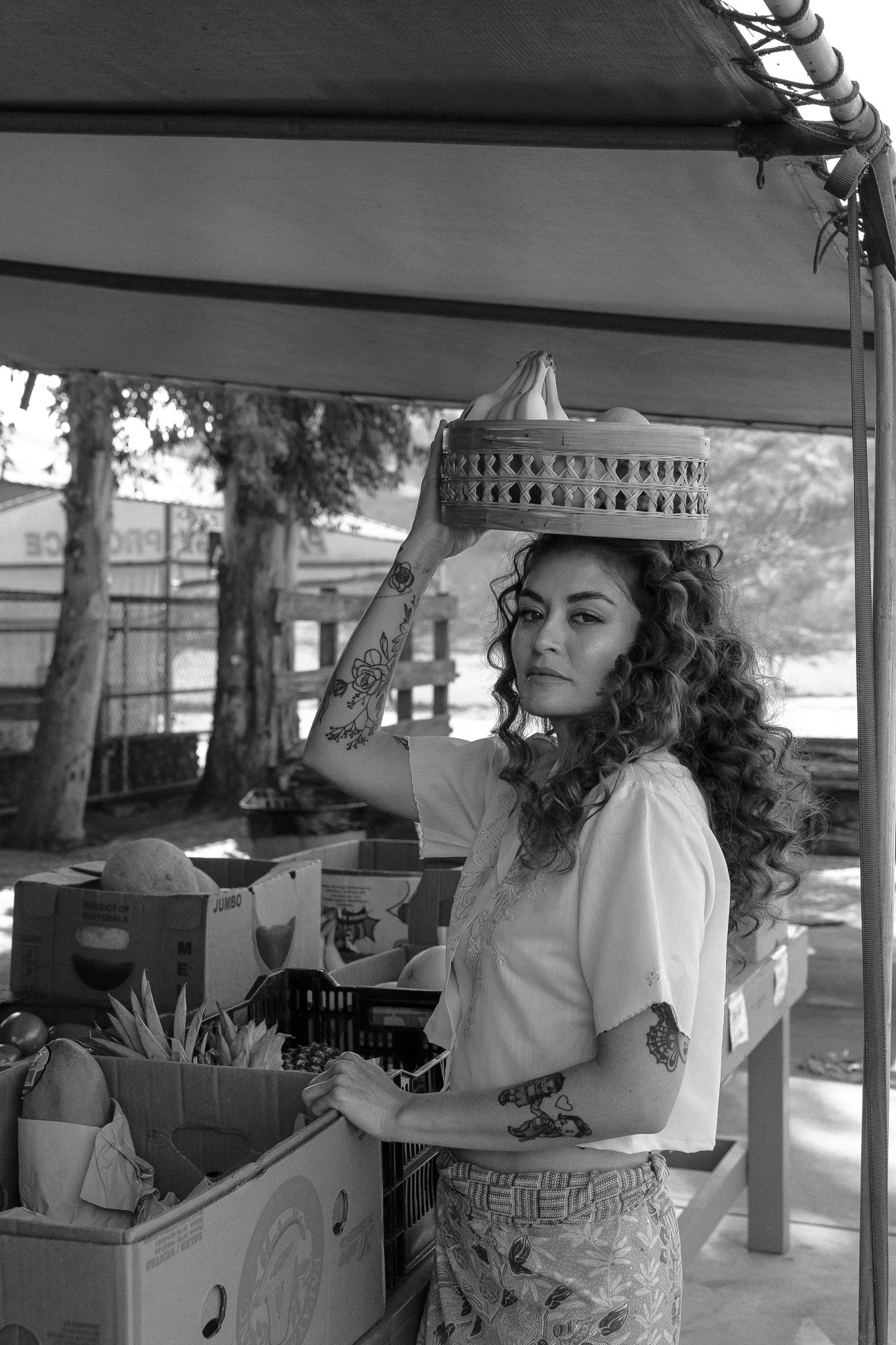 Black and white photo of a woman with curly hair and tattoos holding a basket of fruit on her head at a market stall.