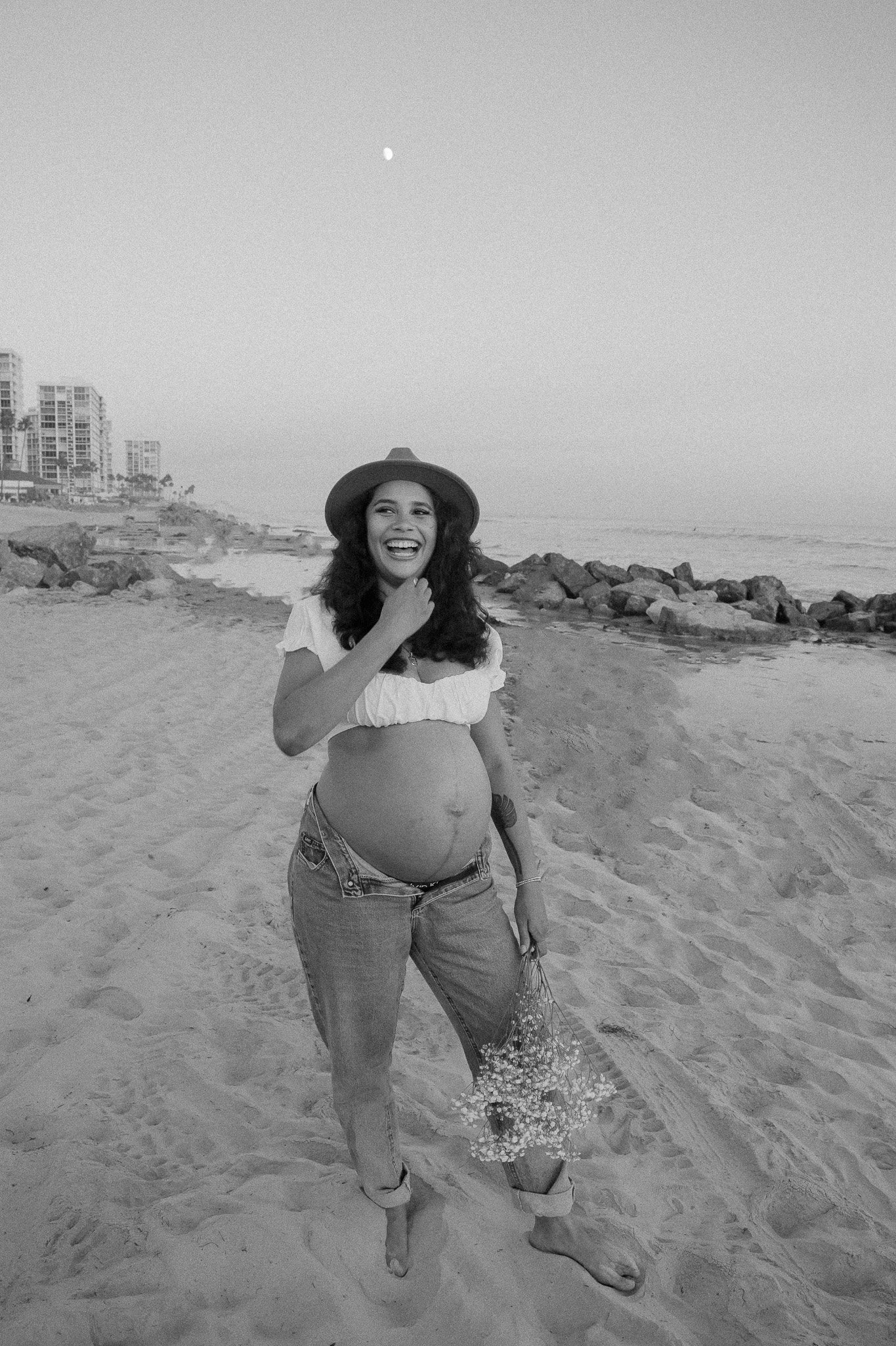 Pregnant woman in jeans and a hat laughing on a beach, holding flowers, with buildings and the ocean in the background.