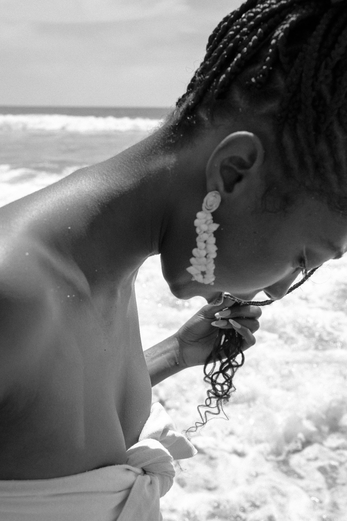 Black and white photo of a person with braided hair at the beach, wearing shell earrings, holding a braid, with waves in the background.