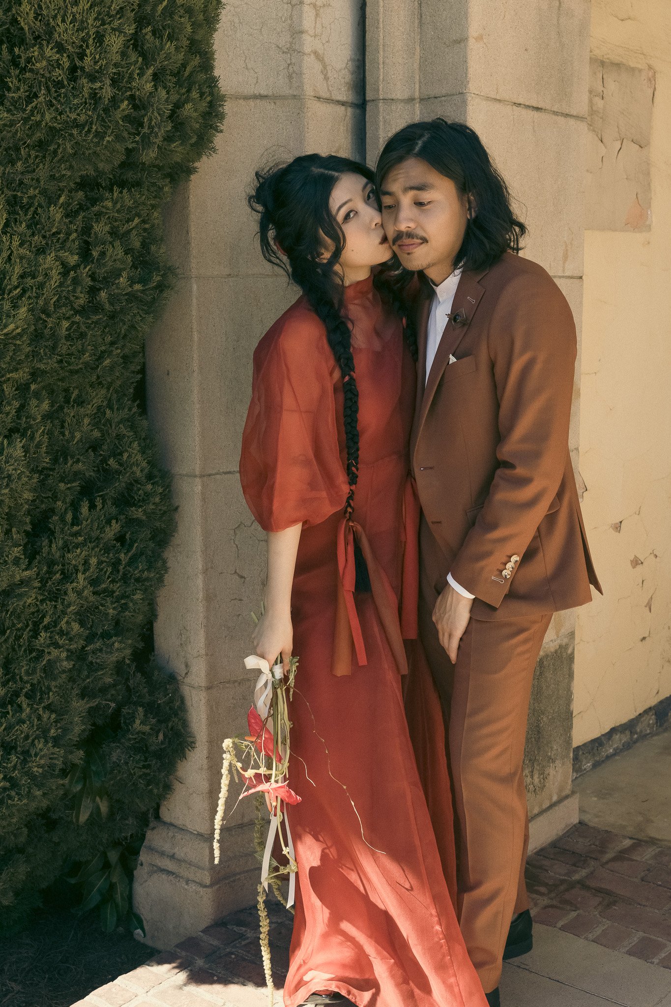 A couple standing close together by a stone wall, dressed in matching reddish attire. The woman, holding a small bouquet, leans in as if to kiss the man. Ivy covers part of the stone wall.
