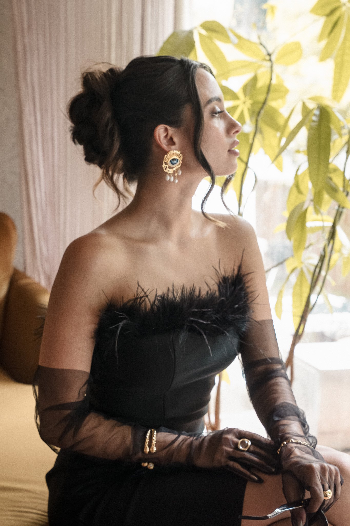 Woman in black feathered dress and sheer gloves, wearing large gold earrings, sitting near a window and greenery.