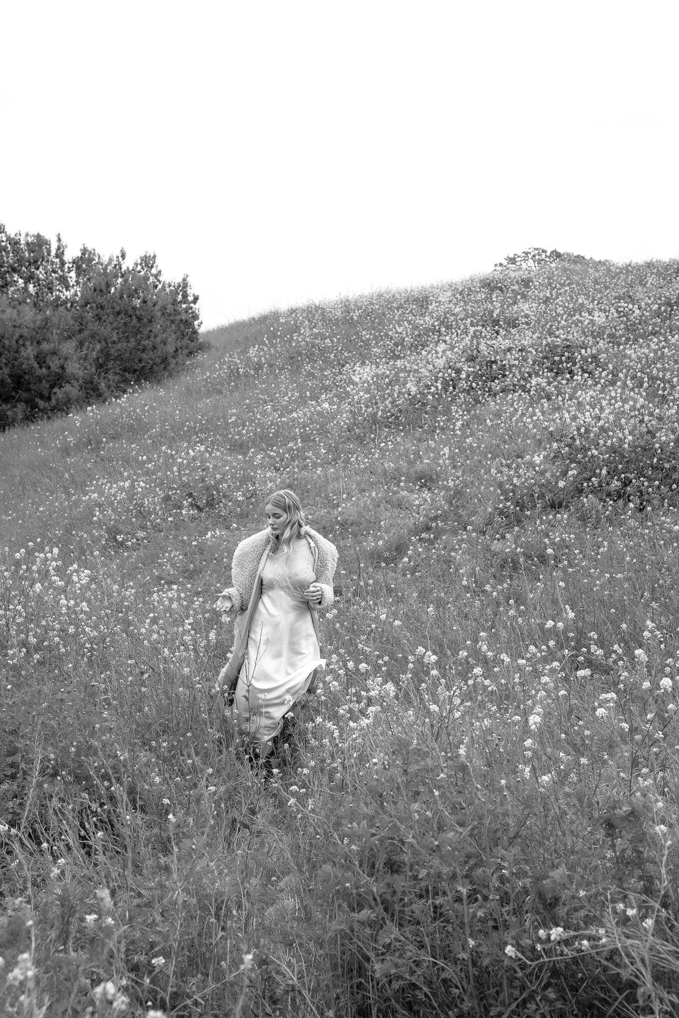 Woman in a field of wildflowers, wearing a long dress and coat, walking on a grassy hillside in black and white.