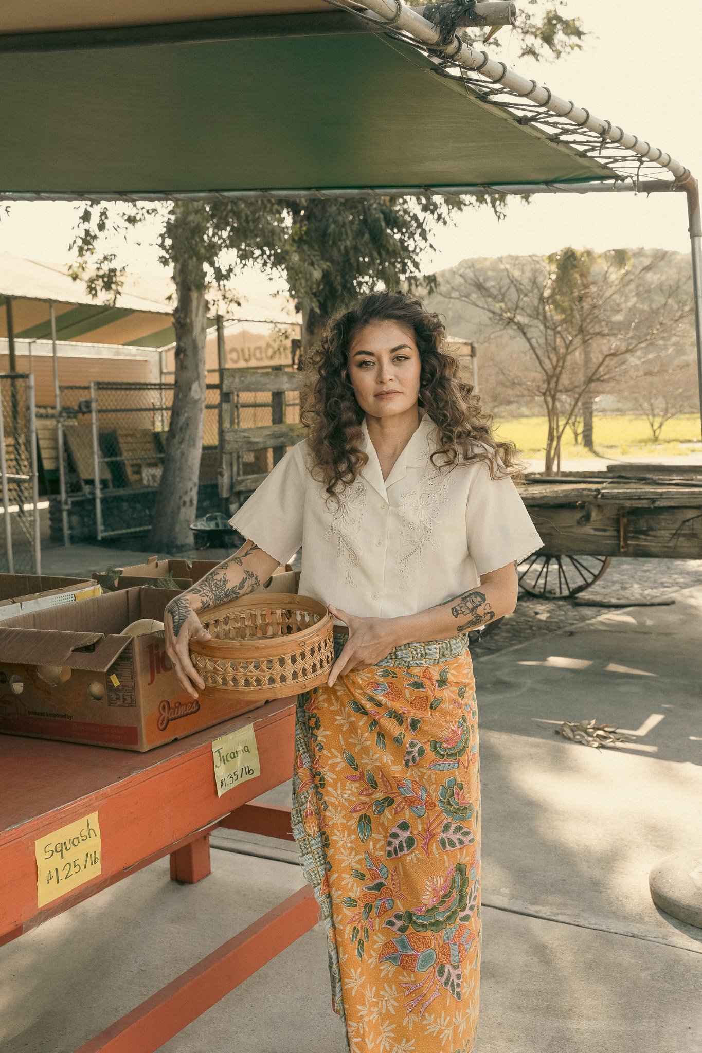Woman holding a basket at an outdoor market stand with boxes labeled "Squash $1.25/lb" and "Tuania $1.35/lb."