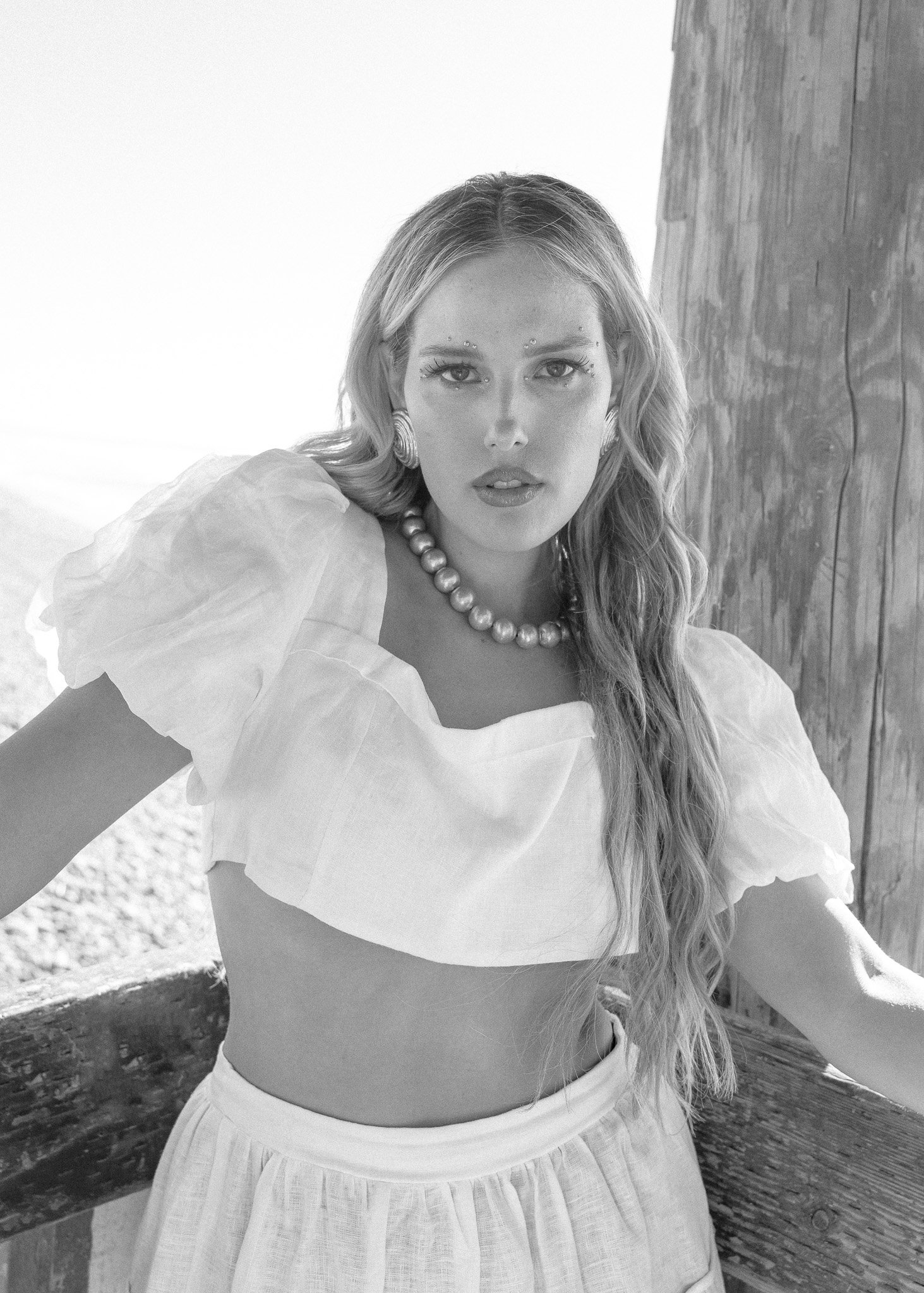 Black and white photo of a woman in a white crop top and skirt, wearing a pearl necklace and earrings, standing by a wooden structure outdoors.