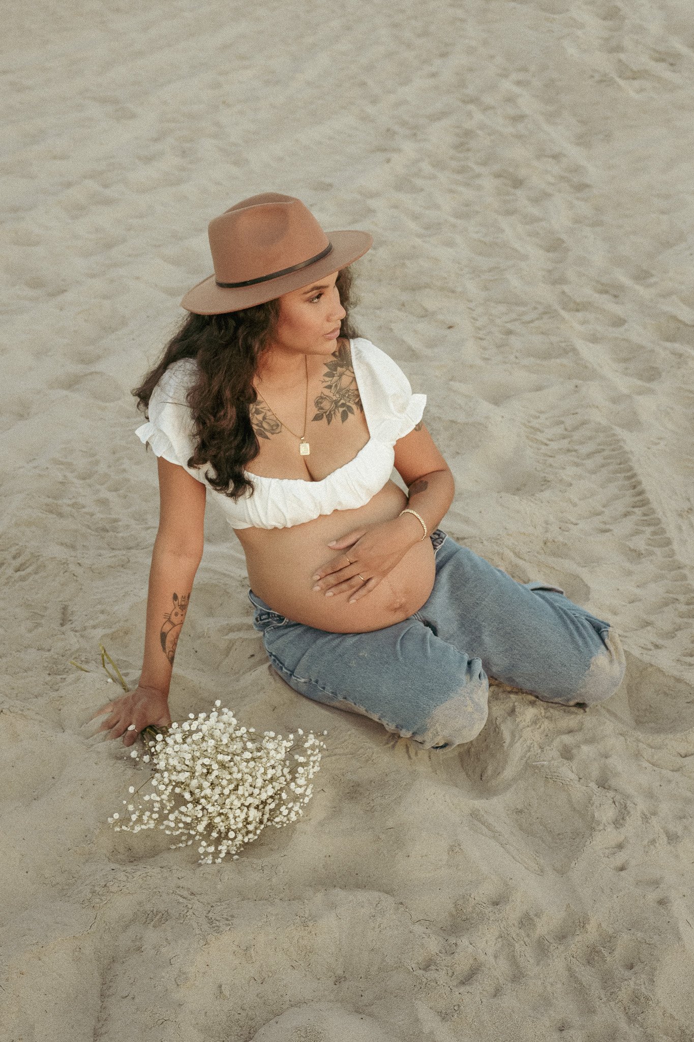 Pregnant woman sitting on sandy beach, wearing a brown hat, white blouse, and jeans, holding her belly. A bouquet of white flowers is beside her.