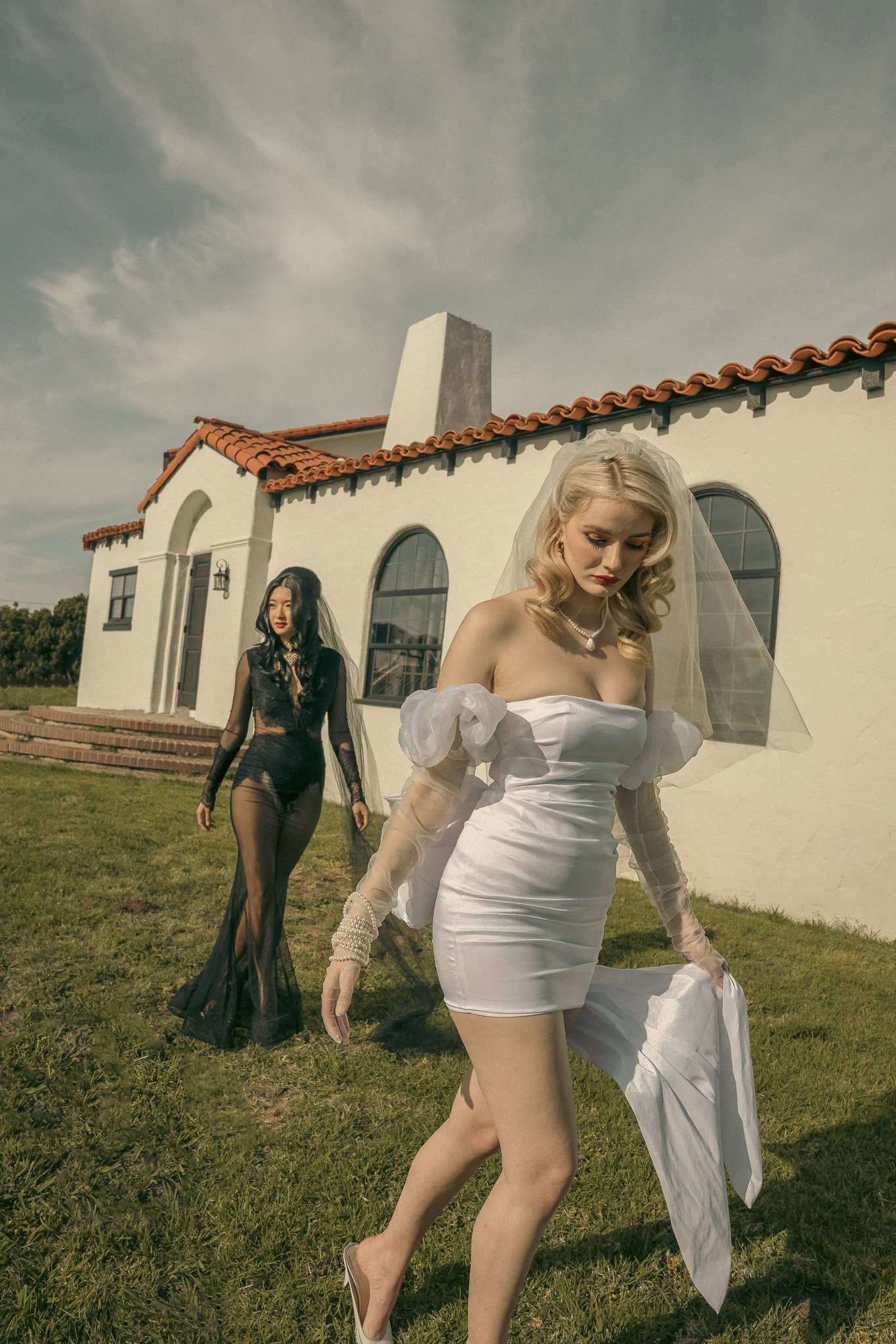 Two women outside a white house with red roof tiles. One wears a short white dress and veil, while the other wears a long black lace dress. Grass lawn in the foreground.