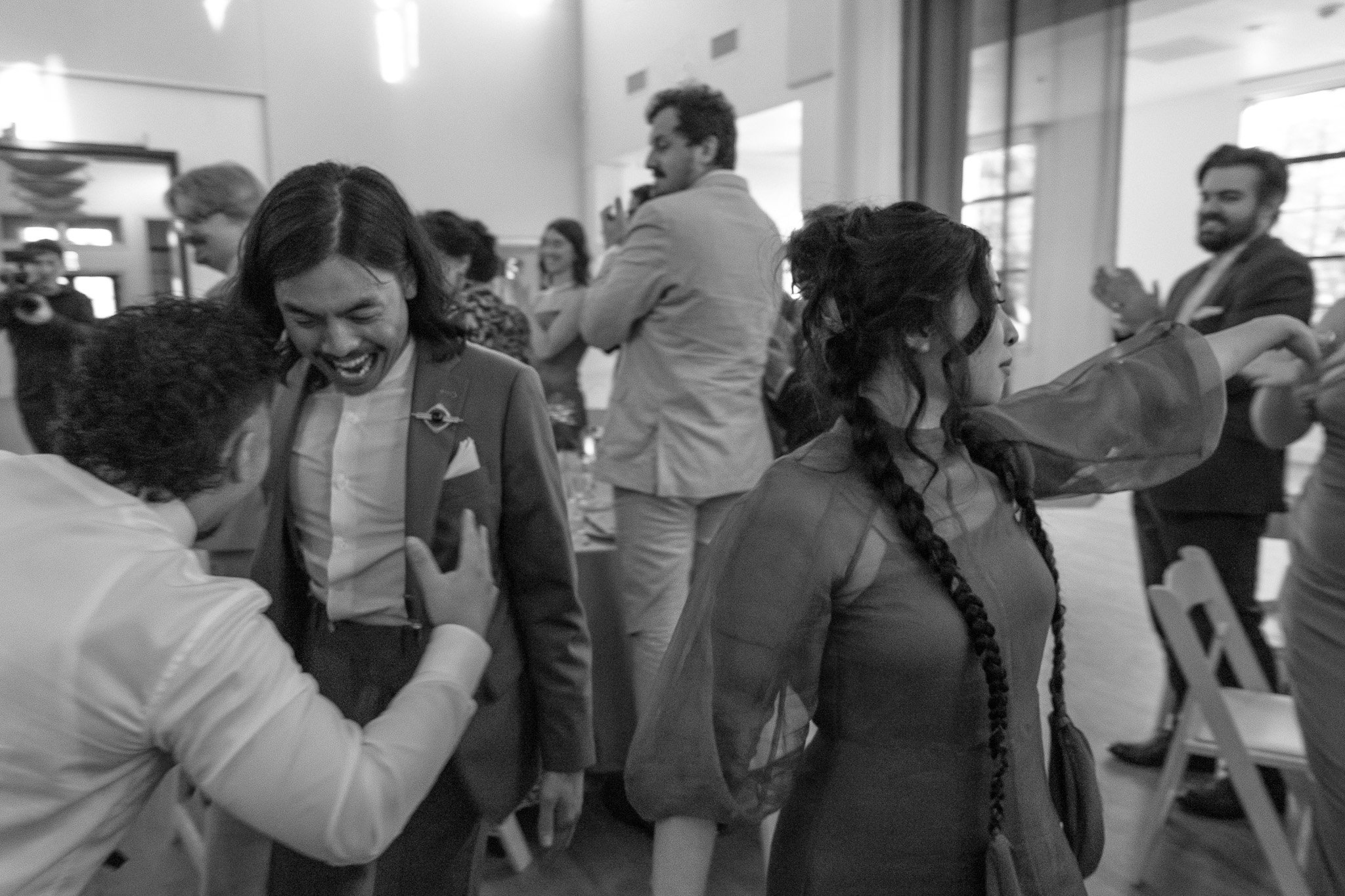 People celebrating at an indoor event, with a man in a suit smiling and interacting with others, and a woman with braided hair dancing. The setting appears to be a social gathering, such as a wedding reception.