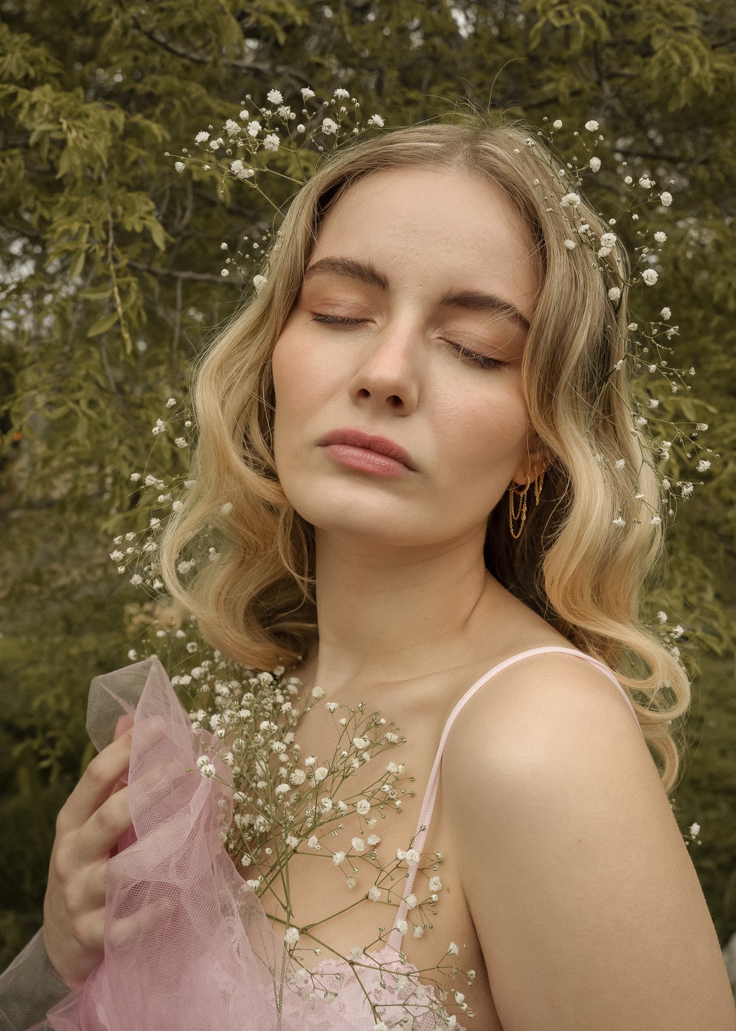 Woman with closed eyes holding flowers, wearing a pink dress, surrounded by greenery.