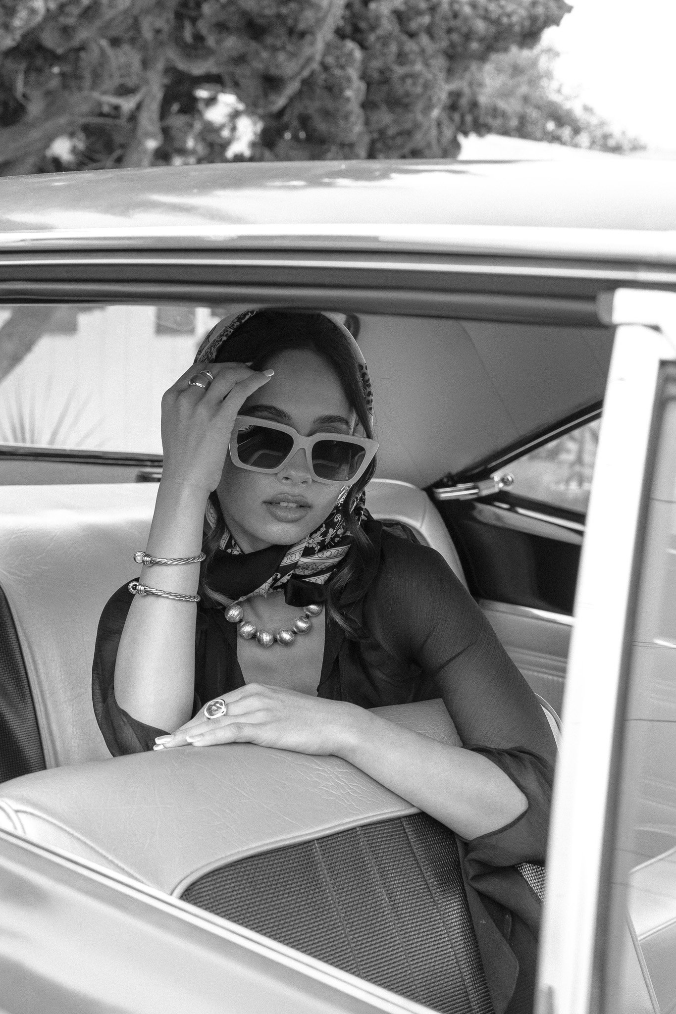 Black and white photo of a woman wearing sunglasses and a headscarf sitting inside a classic car.