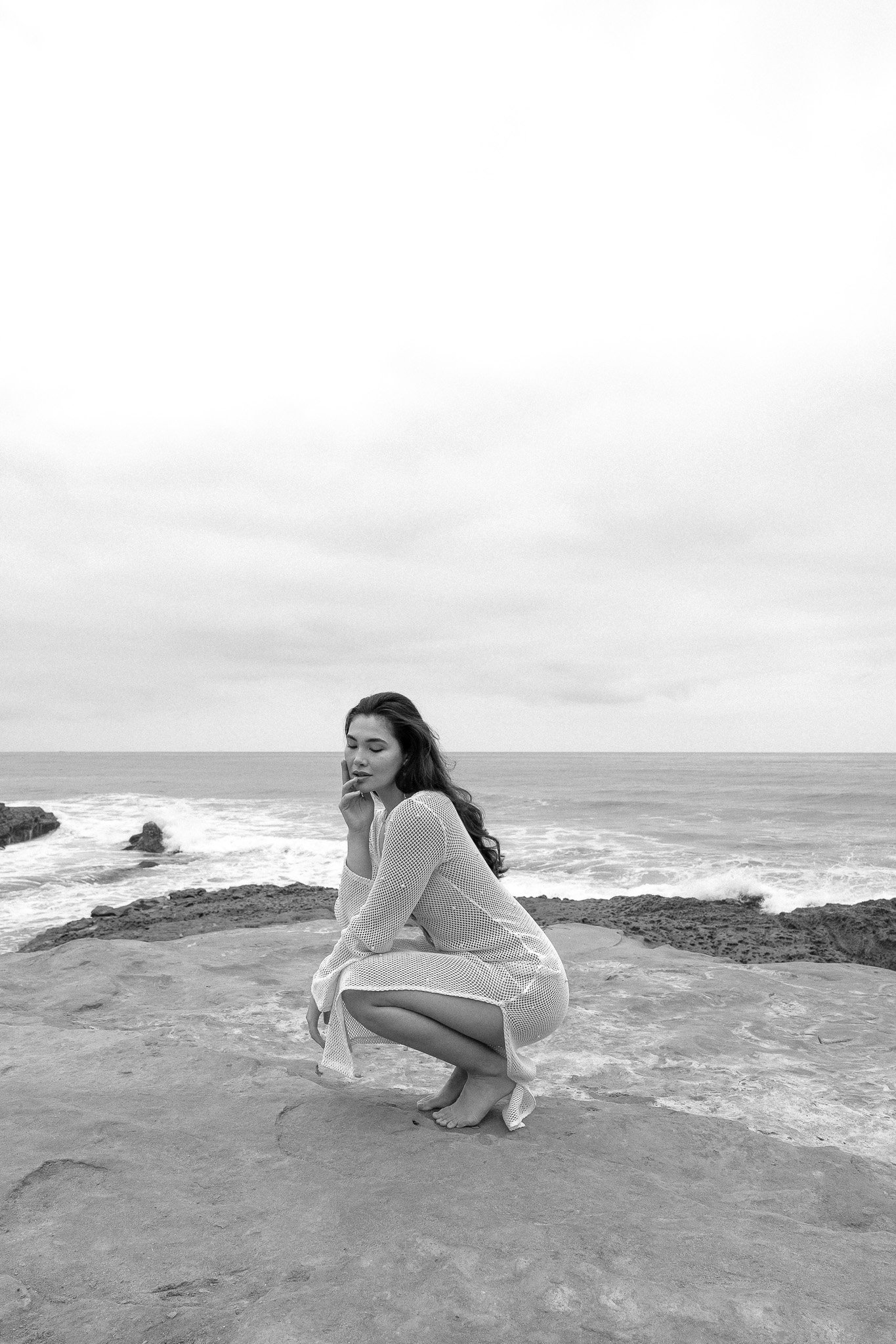 Woman in a sheer dress squatting on a rocky shore with the ocean and cloudy sky in the background, black and white photo.