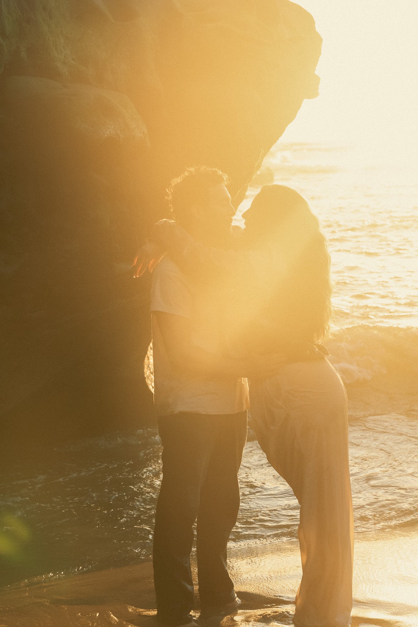 Couple embracing on a beach during sunset, surrounded by golden light.