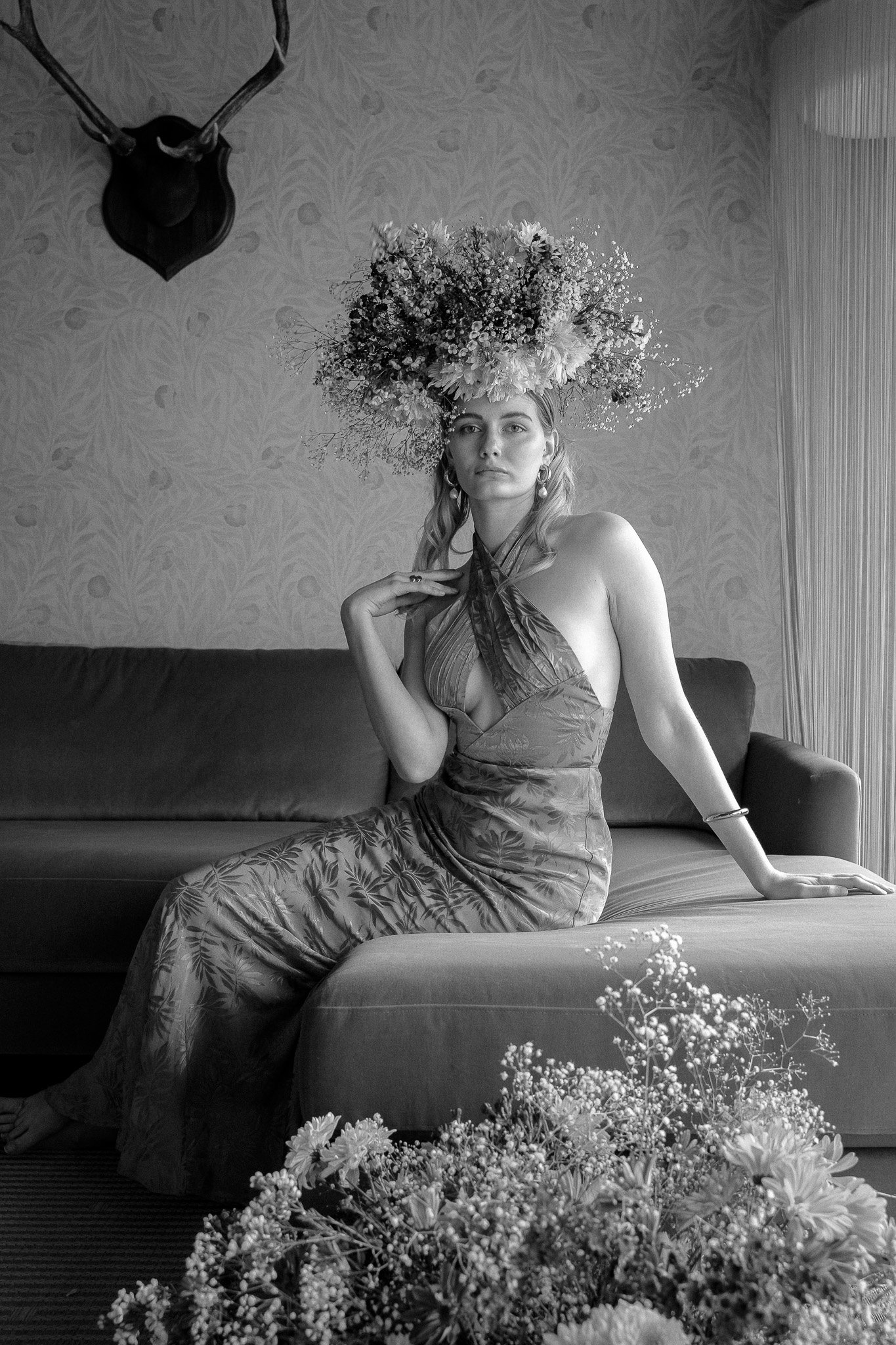 Woman in floral headpiece and dress sitting on a sofa in a room with floral wallpaper and wall-mounted antlers.