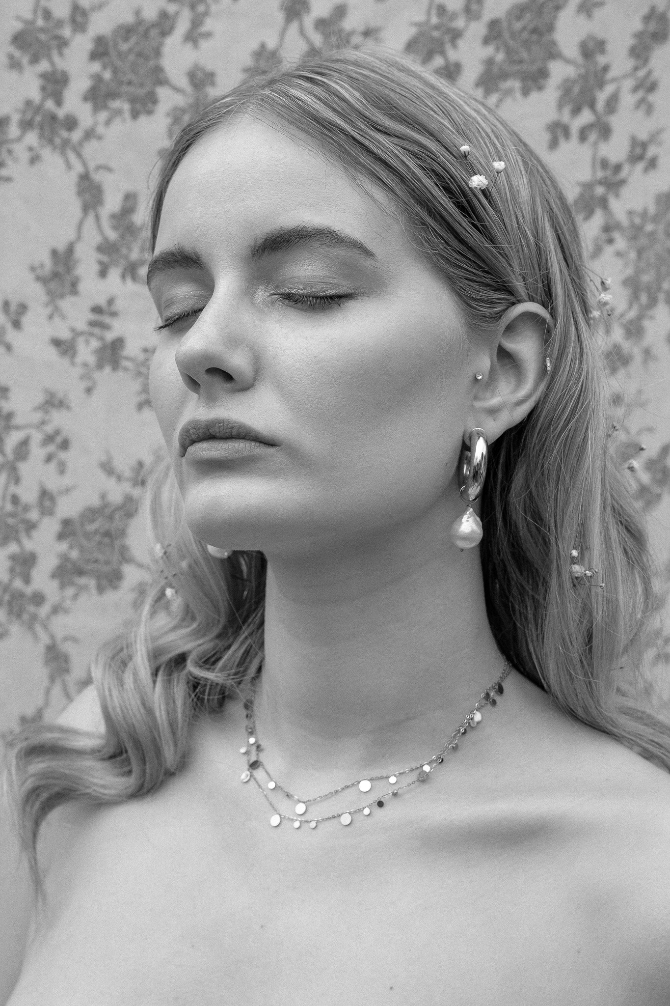 Black and white portrait of a woman with closed eyes, wearing earrings and a delicate necklace against a floral patterned background.