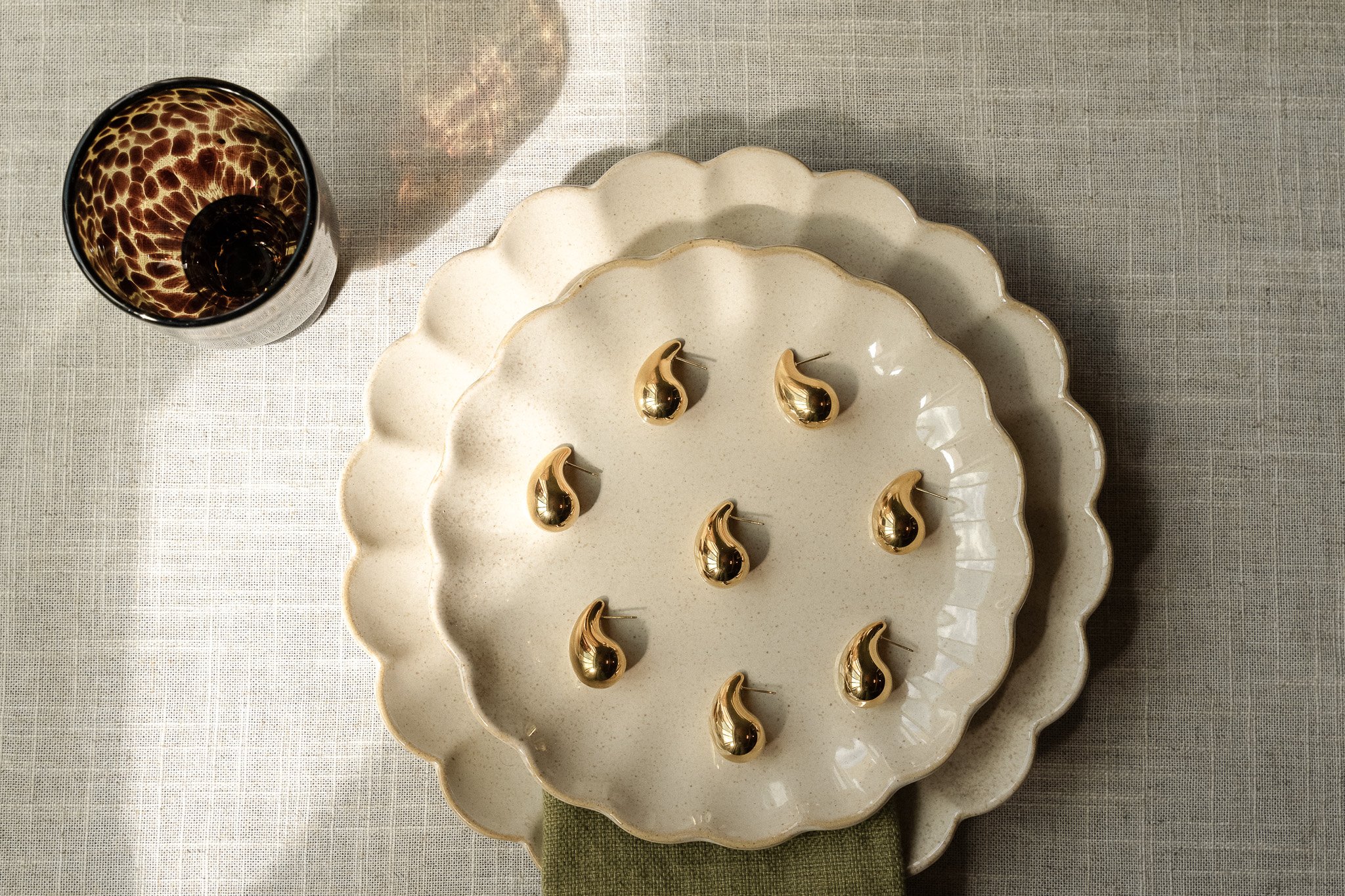 Top view of two beige scalloped plates stacked on a textured surface, adorned with gold droplet-shaped pins, alongside a patterned glass.