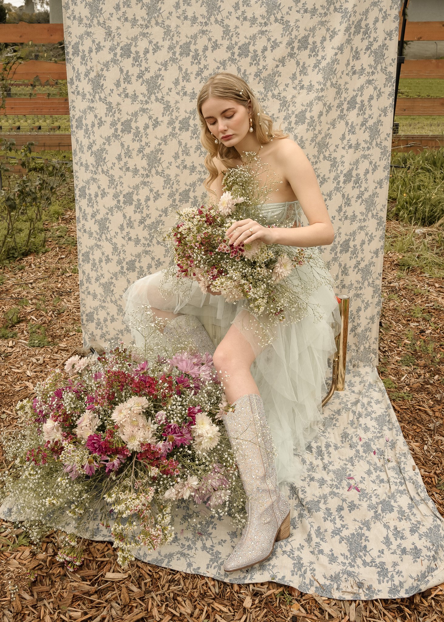 A woman in a light green strapless dress holding a bouquet of flowers, seated against a floral backdrop outdoors. She is wearing glittery boots, surrounded by various flowers and foliage.