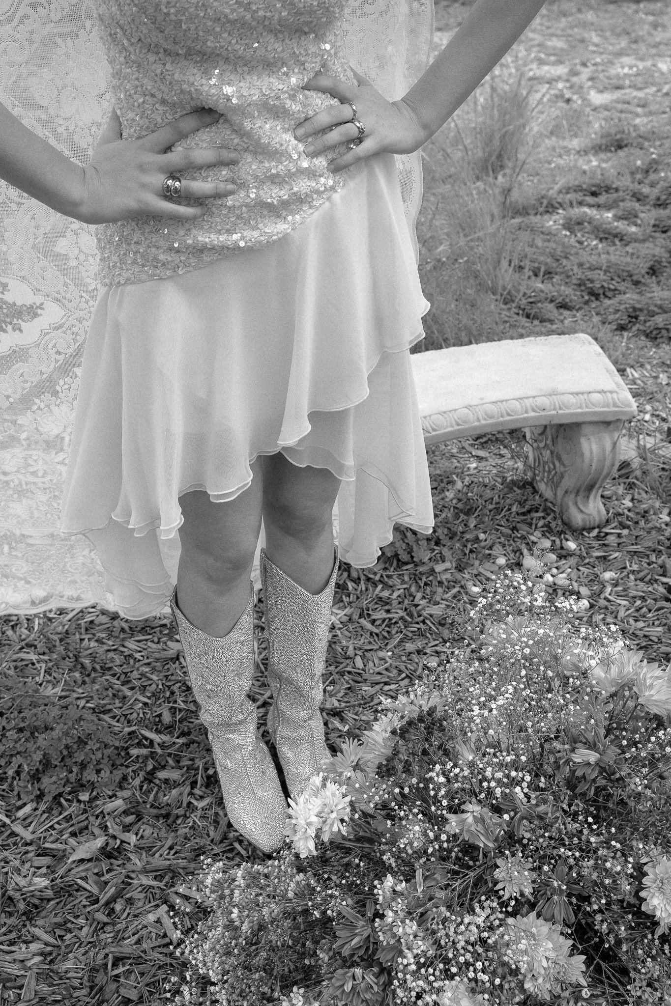 Woman in a sequined dress and glittery boots, standing in a garden with flowers, next to a stone bench.