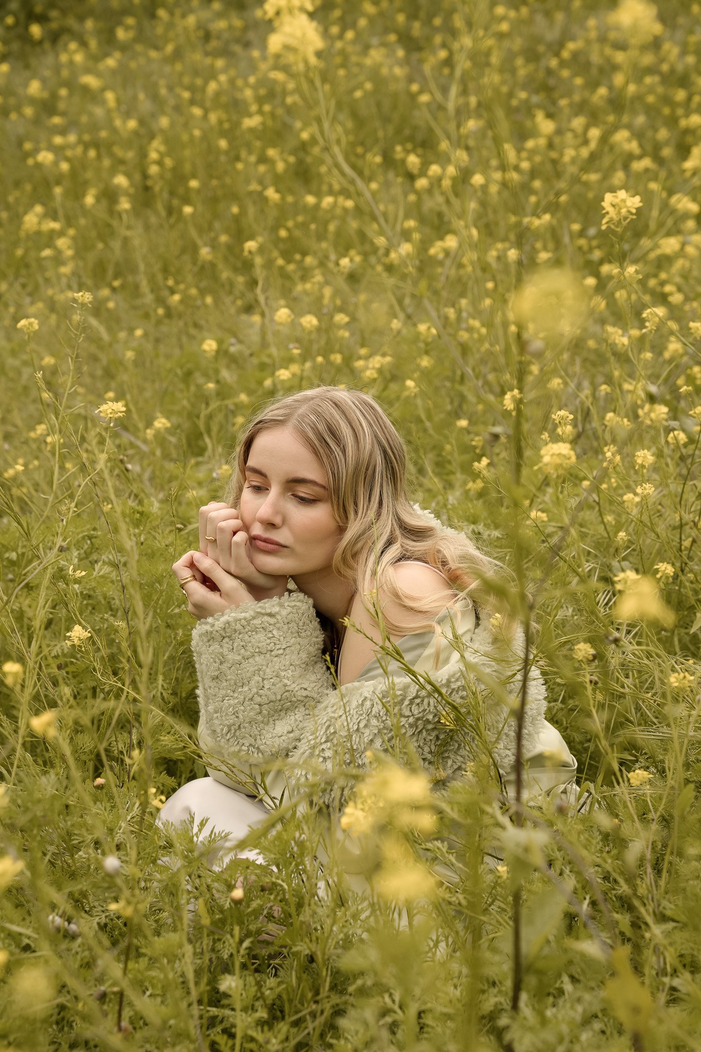 Person with long hair sitting in a field of yellow flowers, wearing a mint green sweater, looking thoughtful.