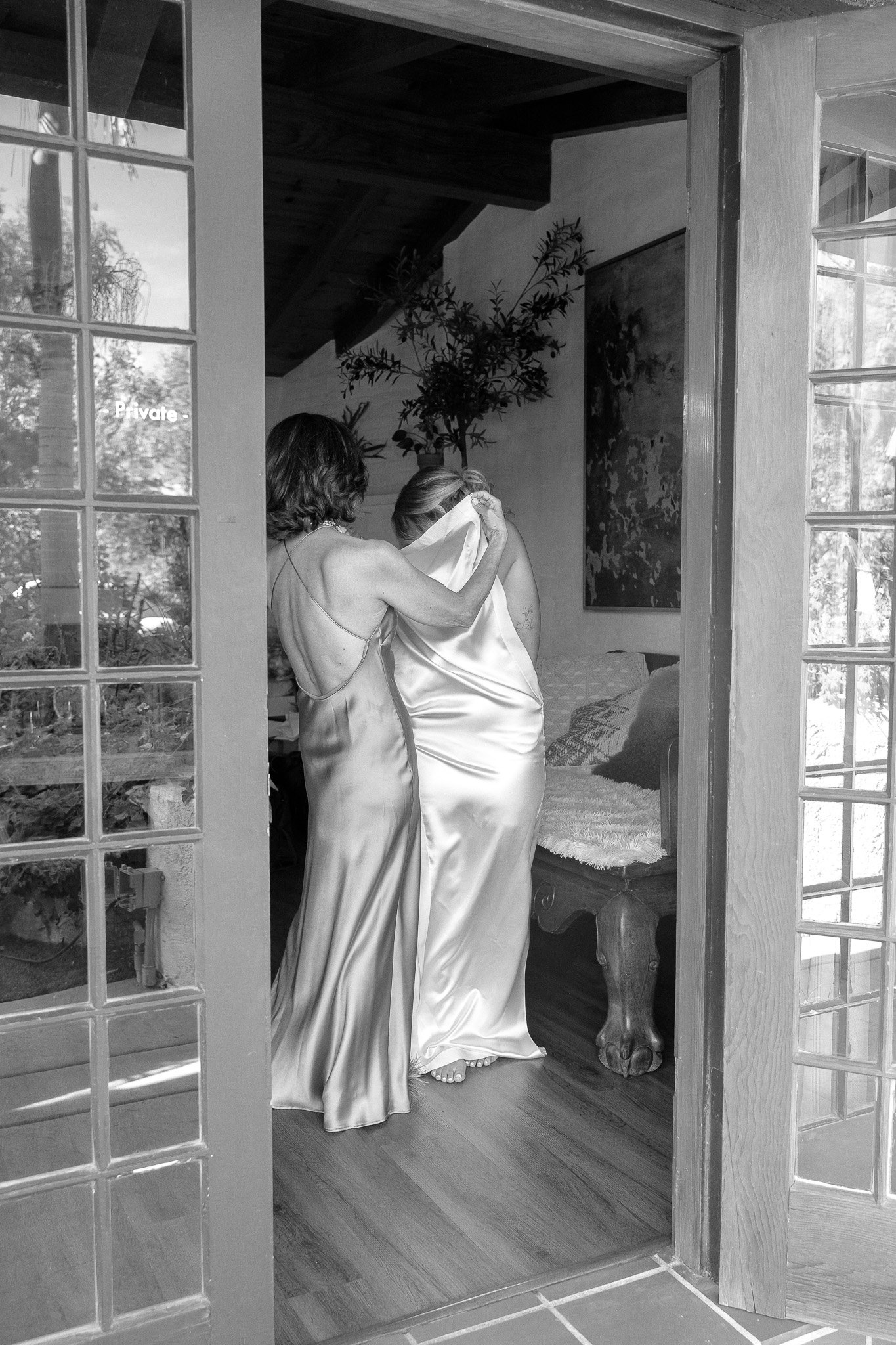 Two women in satin dresses embracing inside a room with a sign on the door reading "Private."