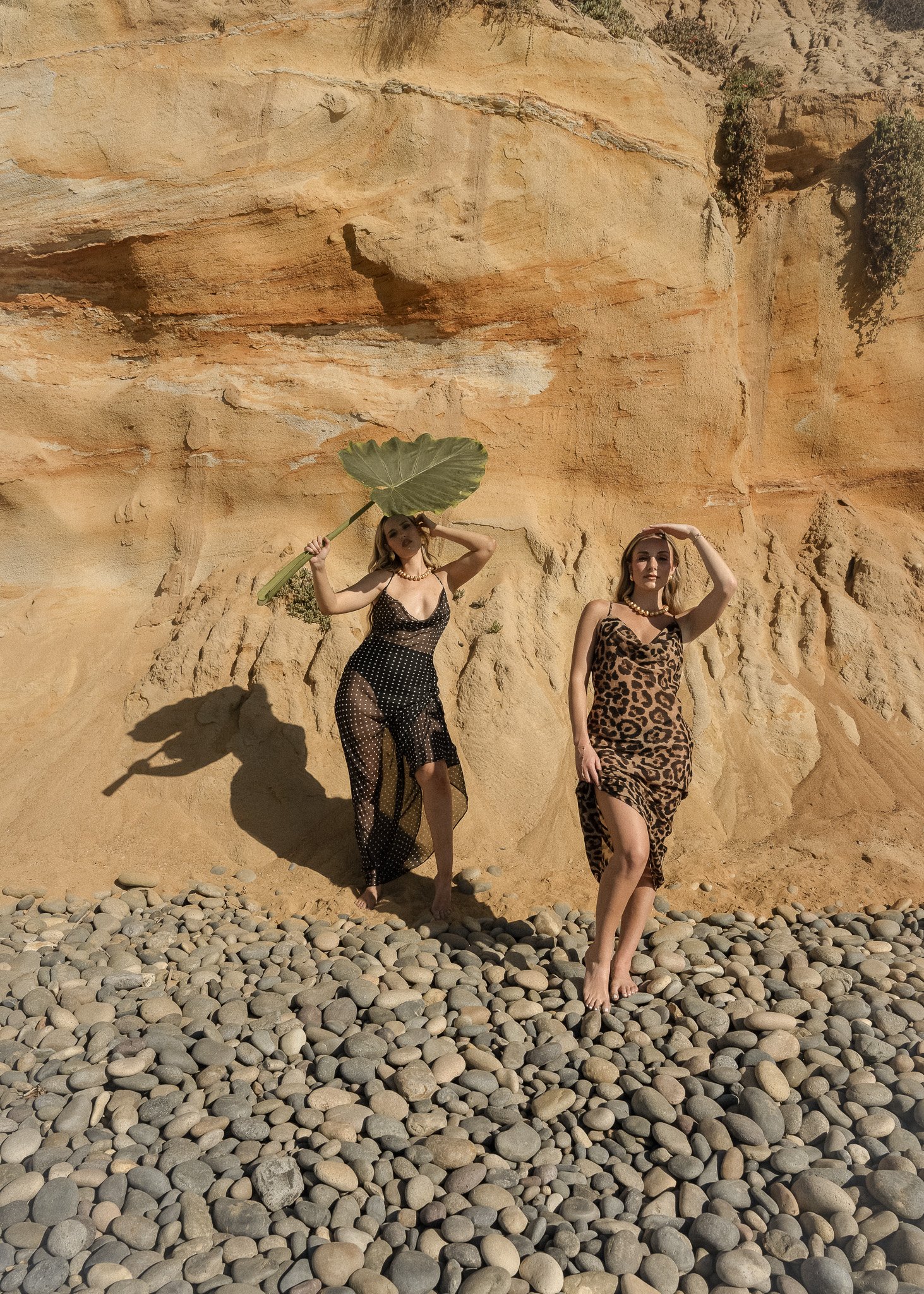 Two women in summer dresses standing on rocky beach with sandstone cliff, one holding large leaf.