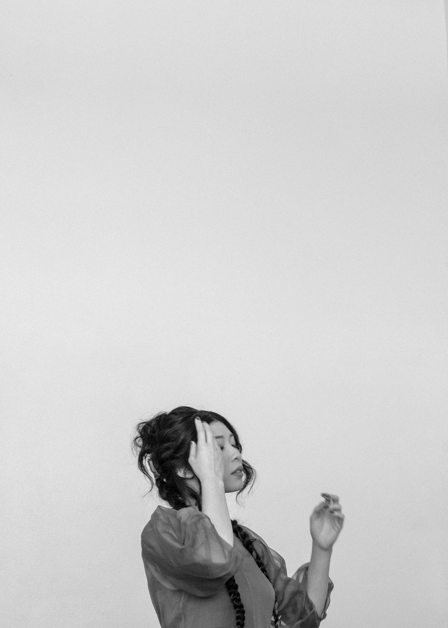 Black and white photo of a woman with long braided hair and a sheer blouse, raising her hands near her head, against a plain background.