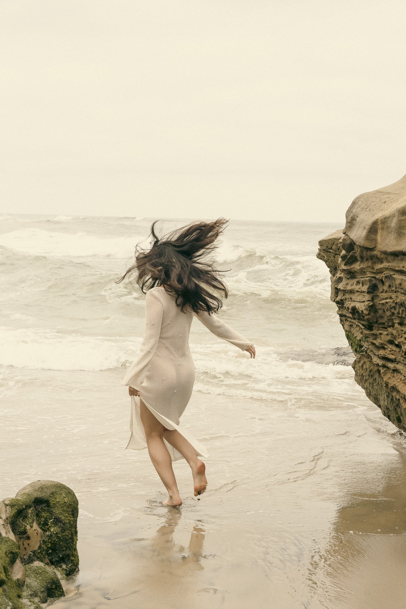 A woman in a white dress with flowing hair standing on a sandy beach near rocky formations, facing the ocean waves.