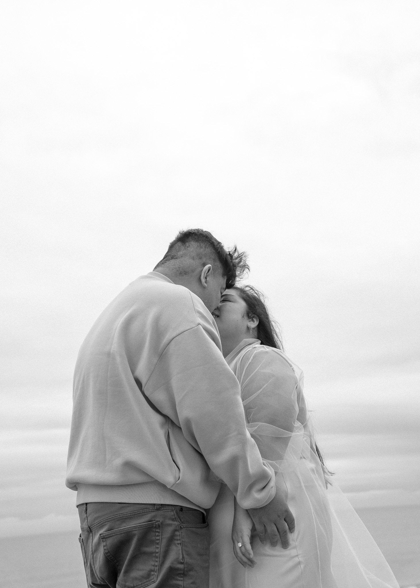 Black and white photo of a couple kissing, with the woman wearing a light, flowing dress and the man in casual clothing against a cloudy sky.