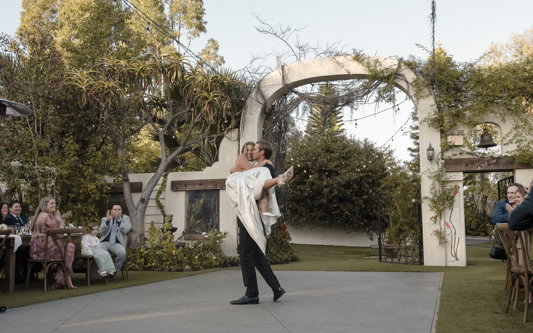 A groom carrying a bride during their wedding celebration, with guests applauding in an outdoor setting with greenery and decorations.