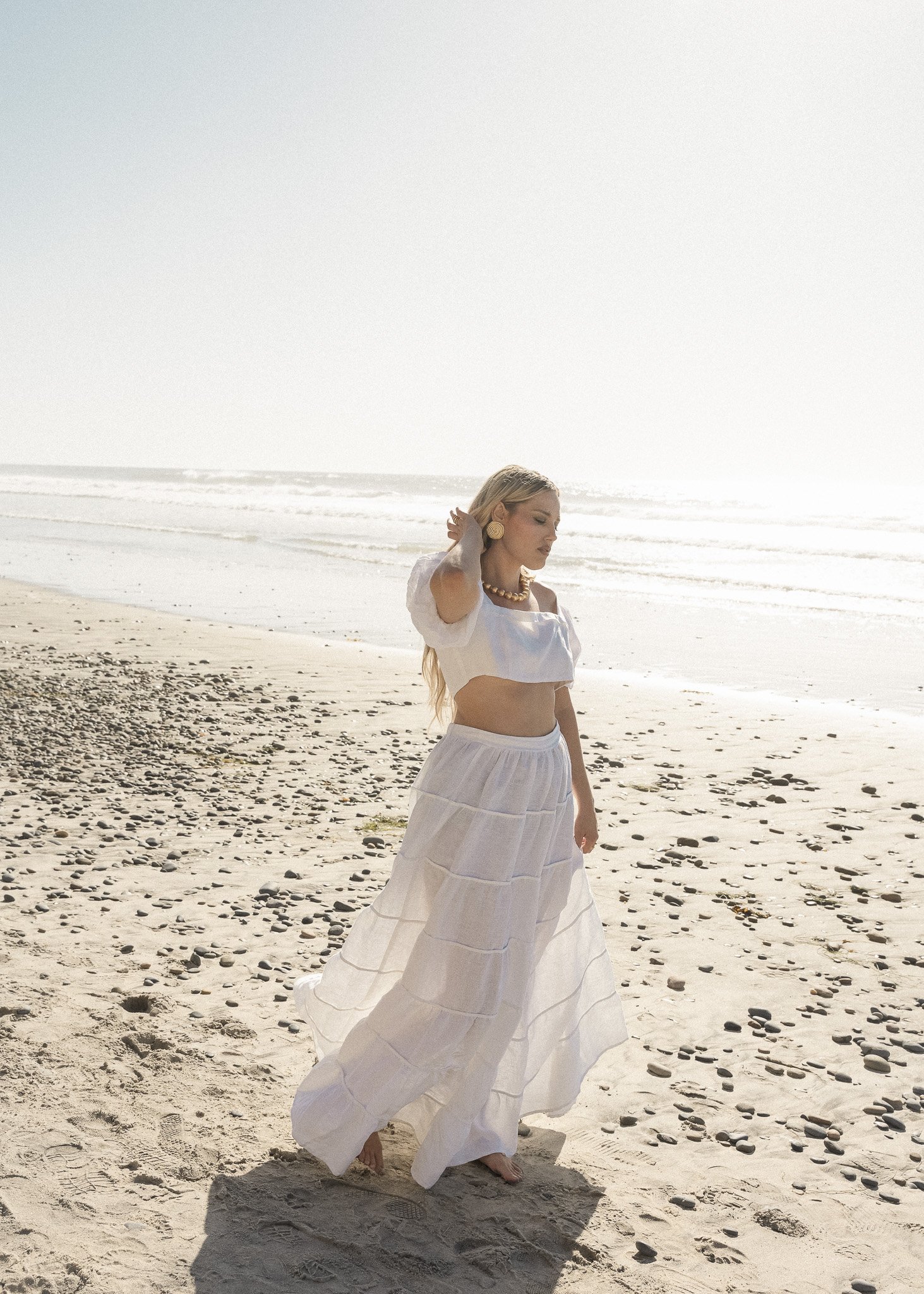 Woman in white outfit walking on a sandy beach with pebbles, ocean in the background.