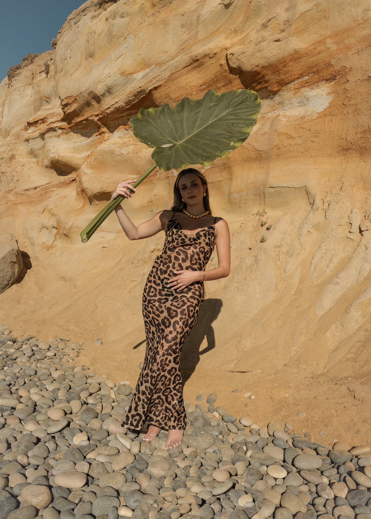 Woman in leopard print dress holding large leaf against rocky cliff.