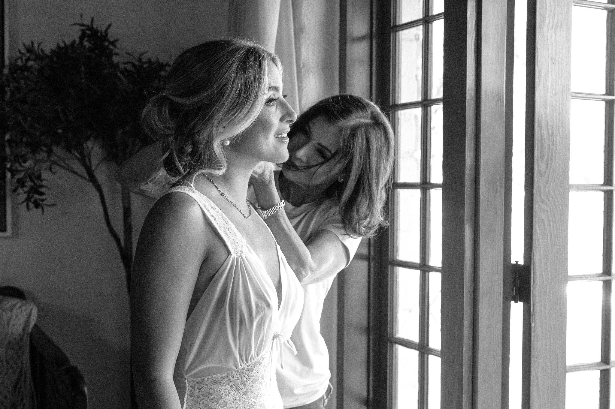 A bride in a lace wedding dress stands by a window smiling, while another woman helps adjust her necklace.