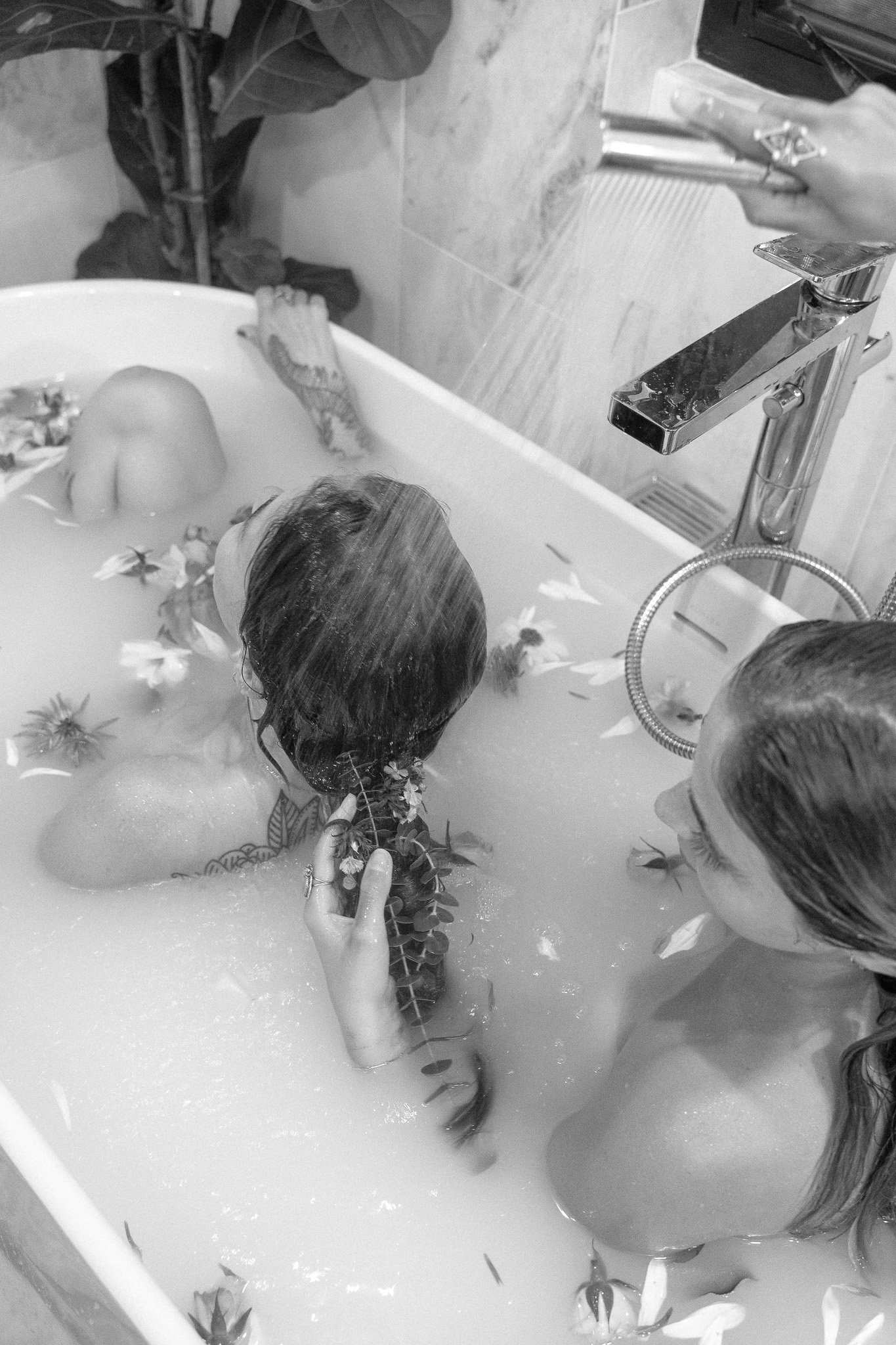 Two people in a bathtub filled with milky water and flower petals, one pouring water on the other's head with a handheld shower. Black and white photograph.