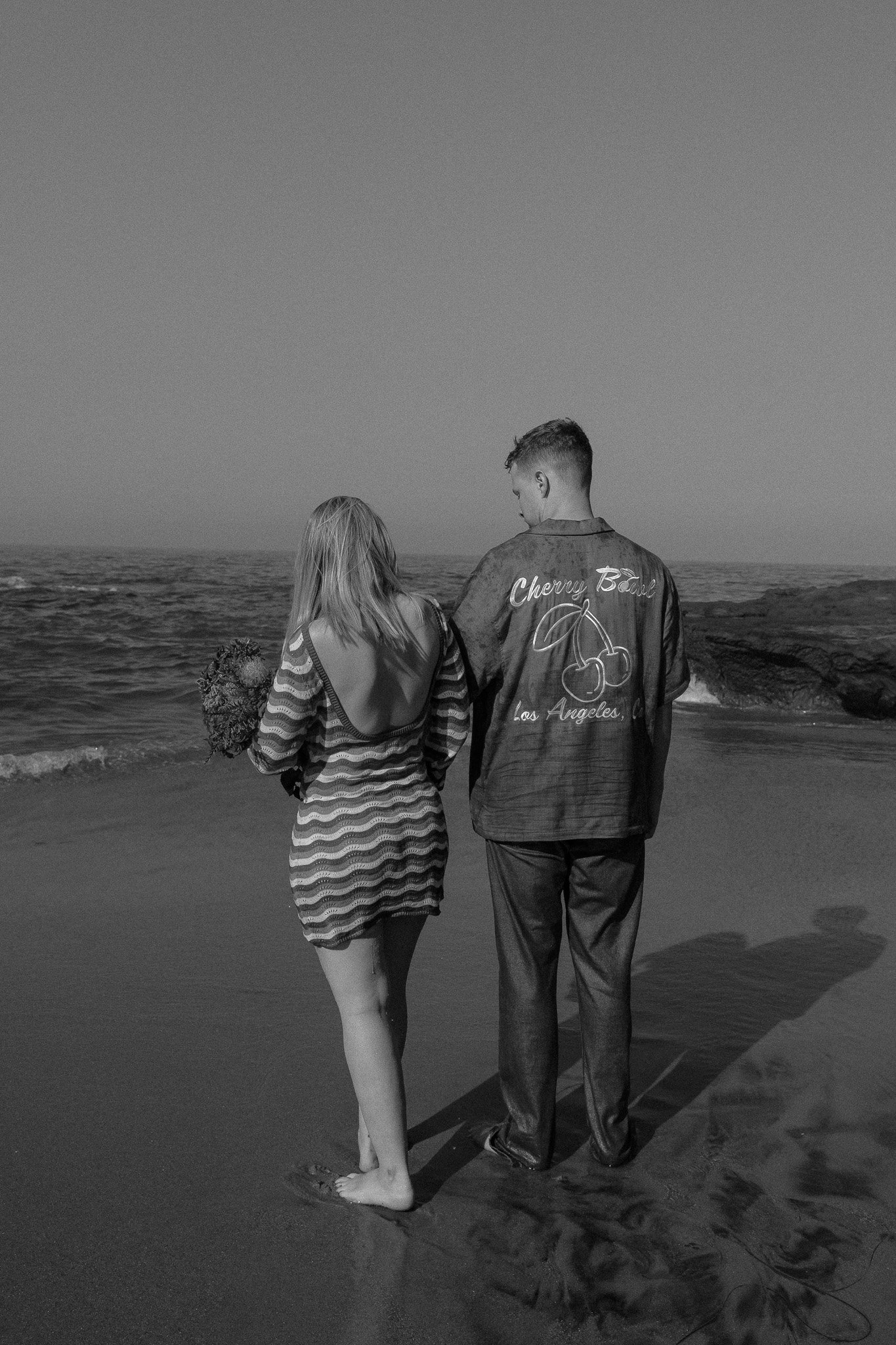 A black and white photo of a man and woman standing on a beach, both facing the ocean. The woman is wearing a striped dress and holding a bouquet. The man is wearing a jacket with the words "Cherry Bomb Los Angeles" on the back, along with a cherry g