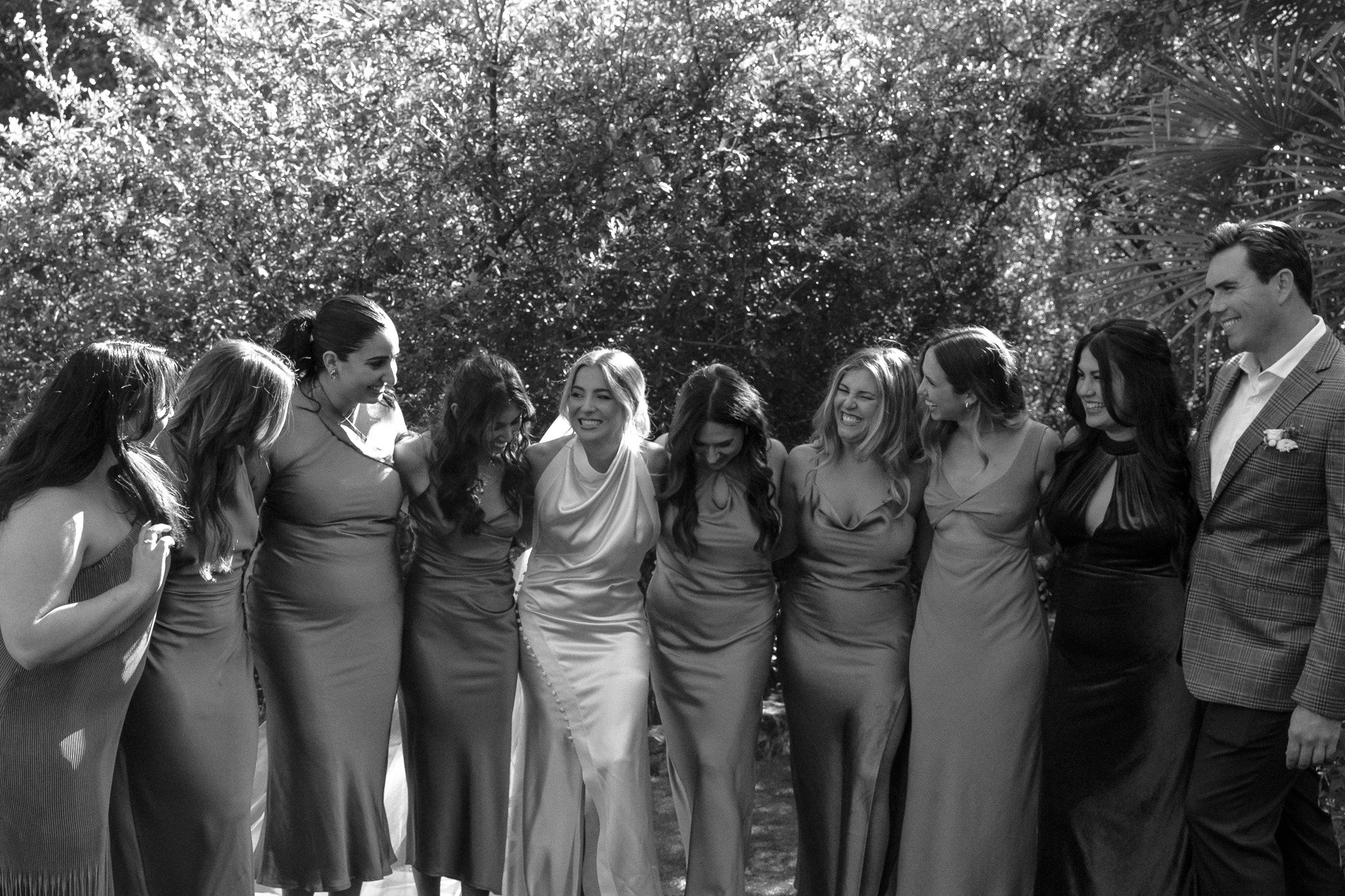 A group of people dressed in formal attire, including a bride in a wedding gown, standing outdoors. They appear to be smiling and enjoying a celebratory moment.