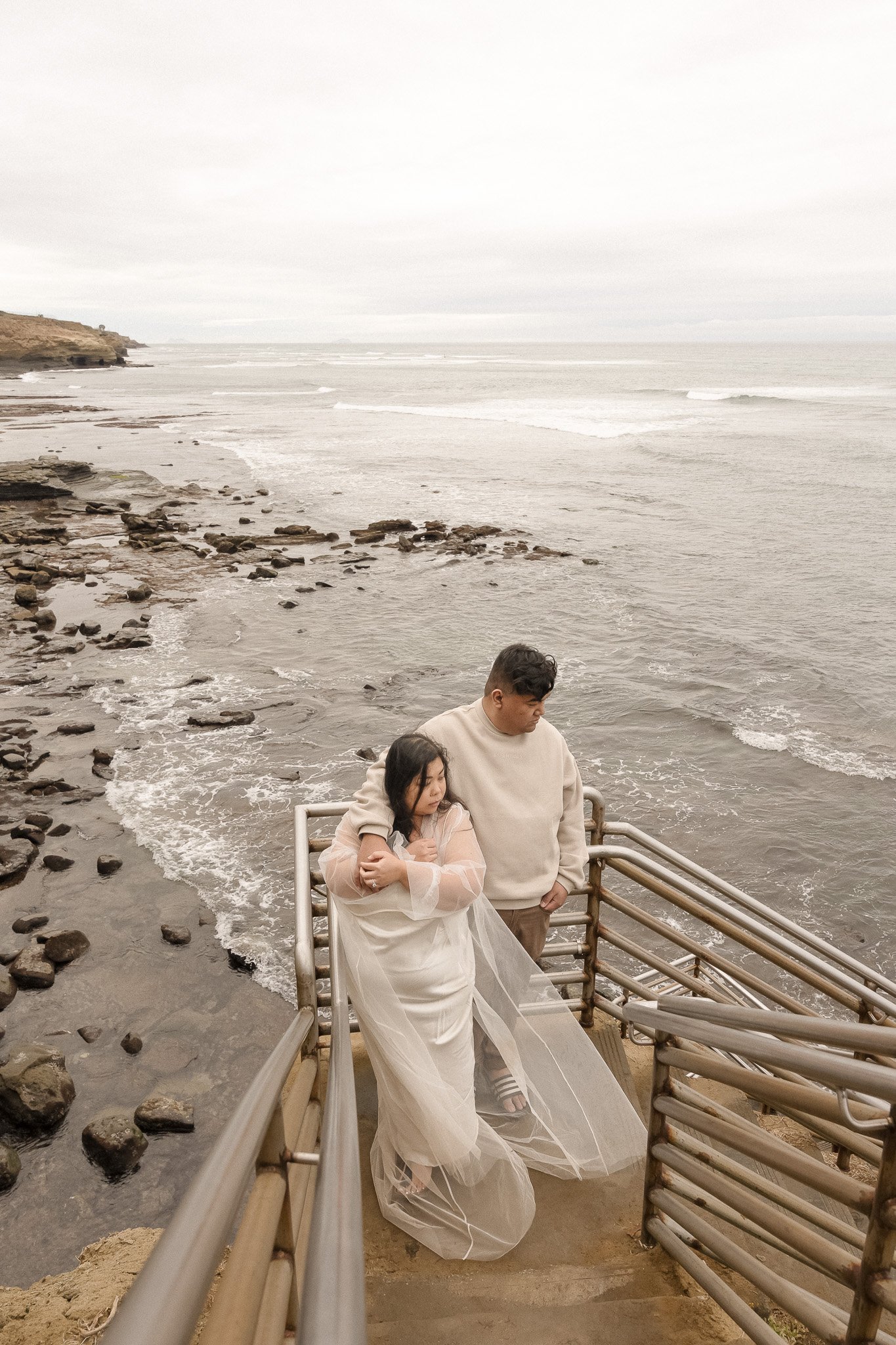 A couple standing on a staircase overlooking a rocky beach and ocean waves. The woman is wearing a white dress with a sheer overlay, and the man is wearing a light-colored sweater. The sky is overcast.