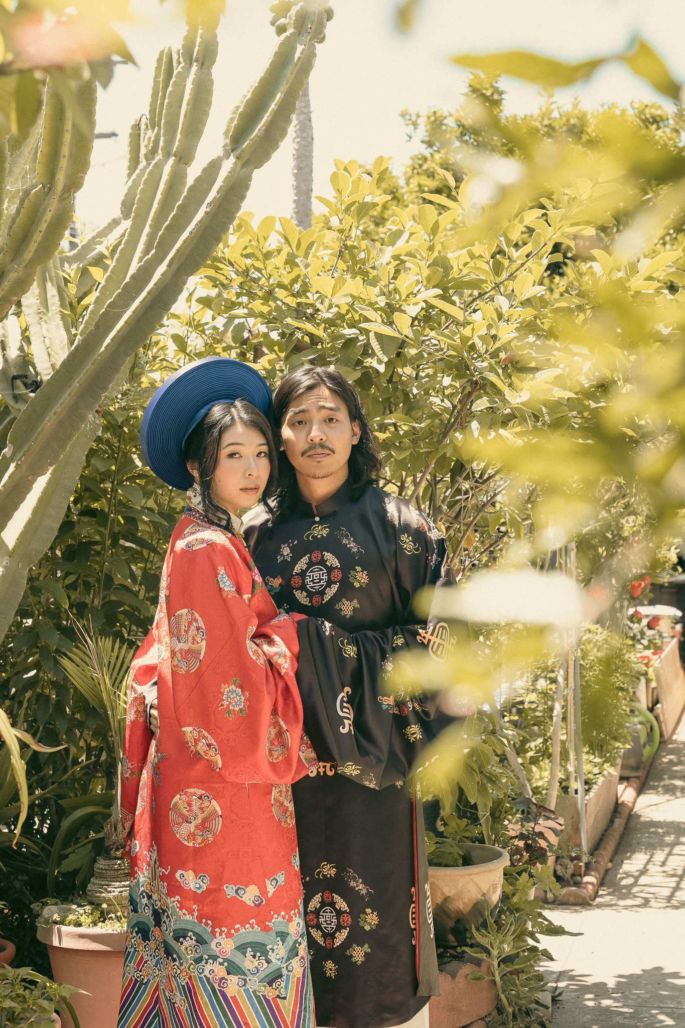 A couple in traditional Asian attire standing outdoors surrounded by plants and trees, with one person wearing a blue hat and a red embroidered garment, and the other in a black outfit with gold embroidery.
