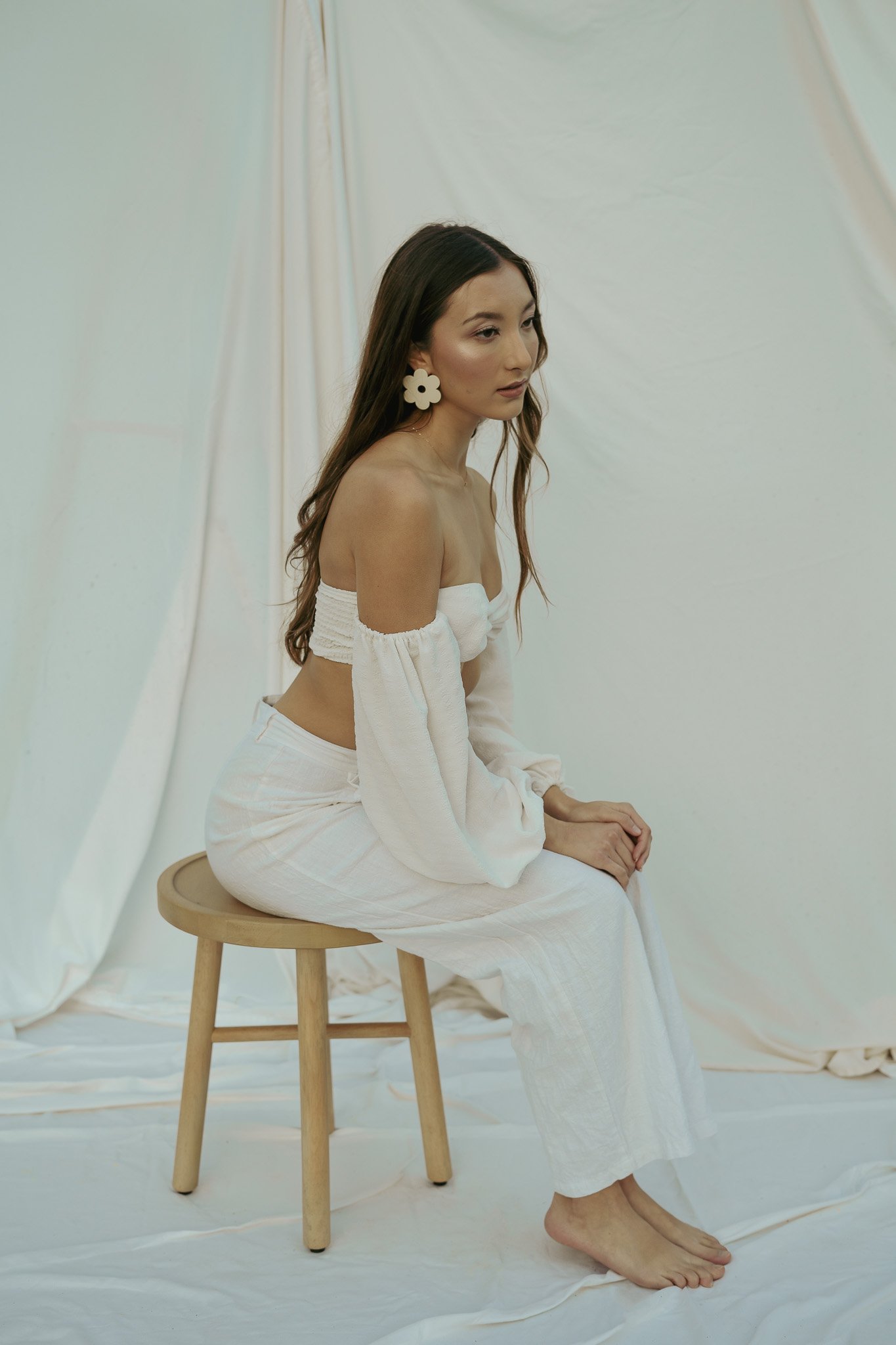 Woman in white off-the-shoulder outfit sitting on wooden stool, posing against a light fabric backdrop.