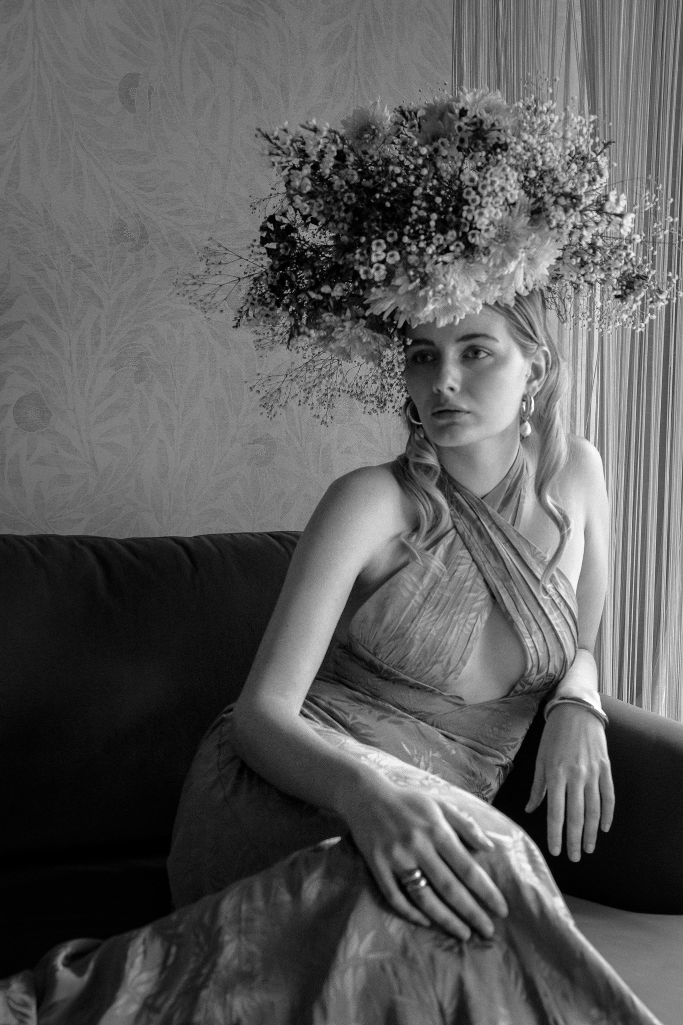 Black and white photo of a woman wearing a large floral headdress and elegant gown, sitting on a couch.