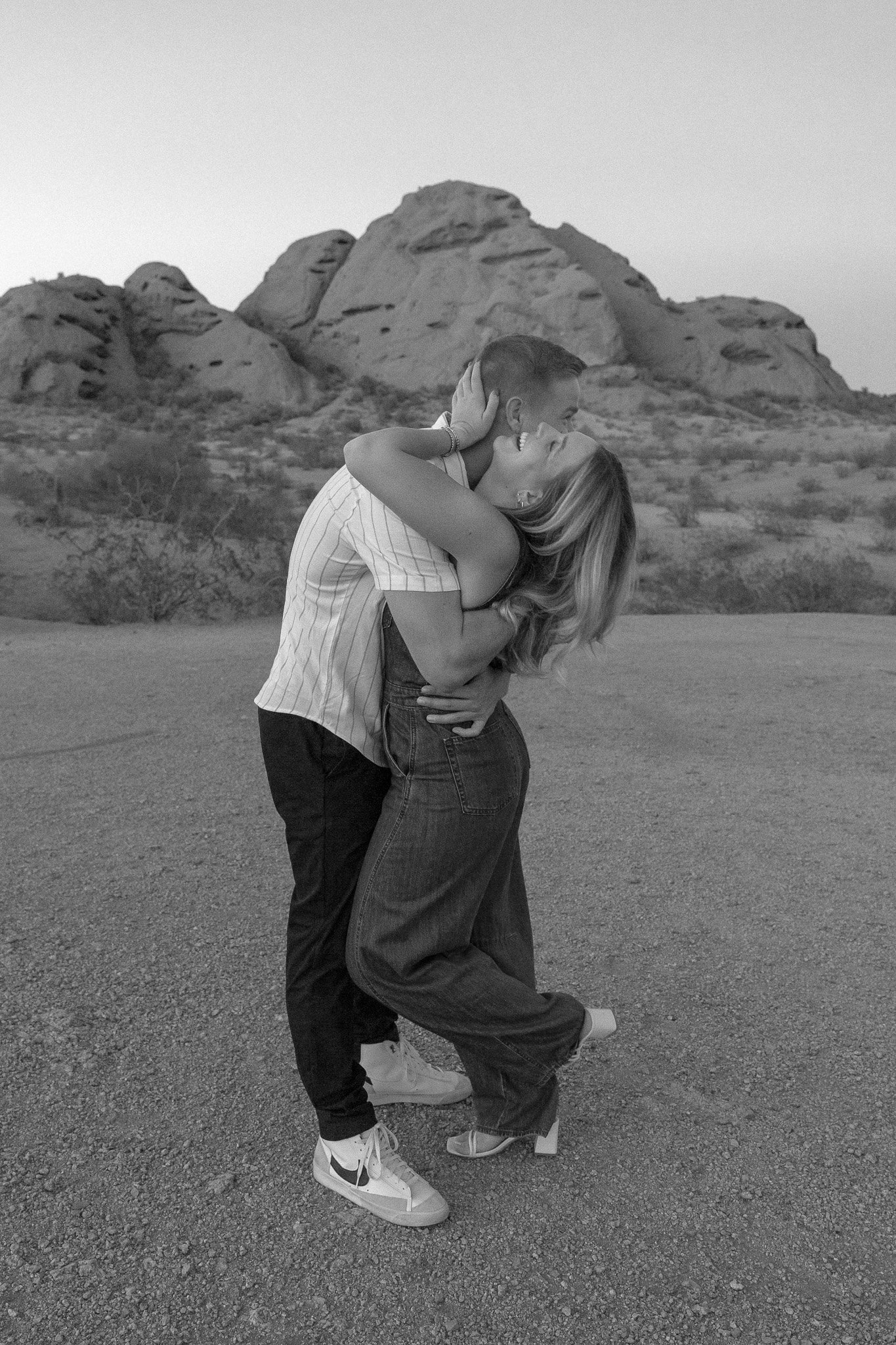 Black and white photo of a couple embracing in a desert landscape, with rocky hills in the background.