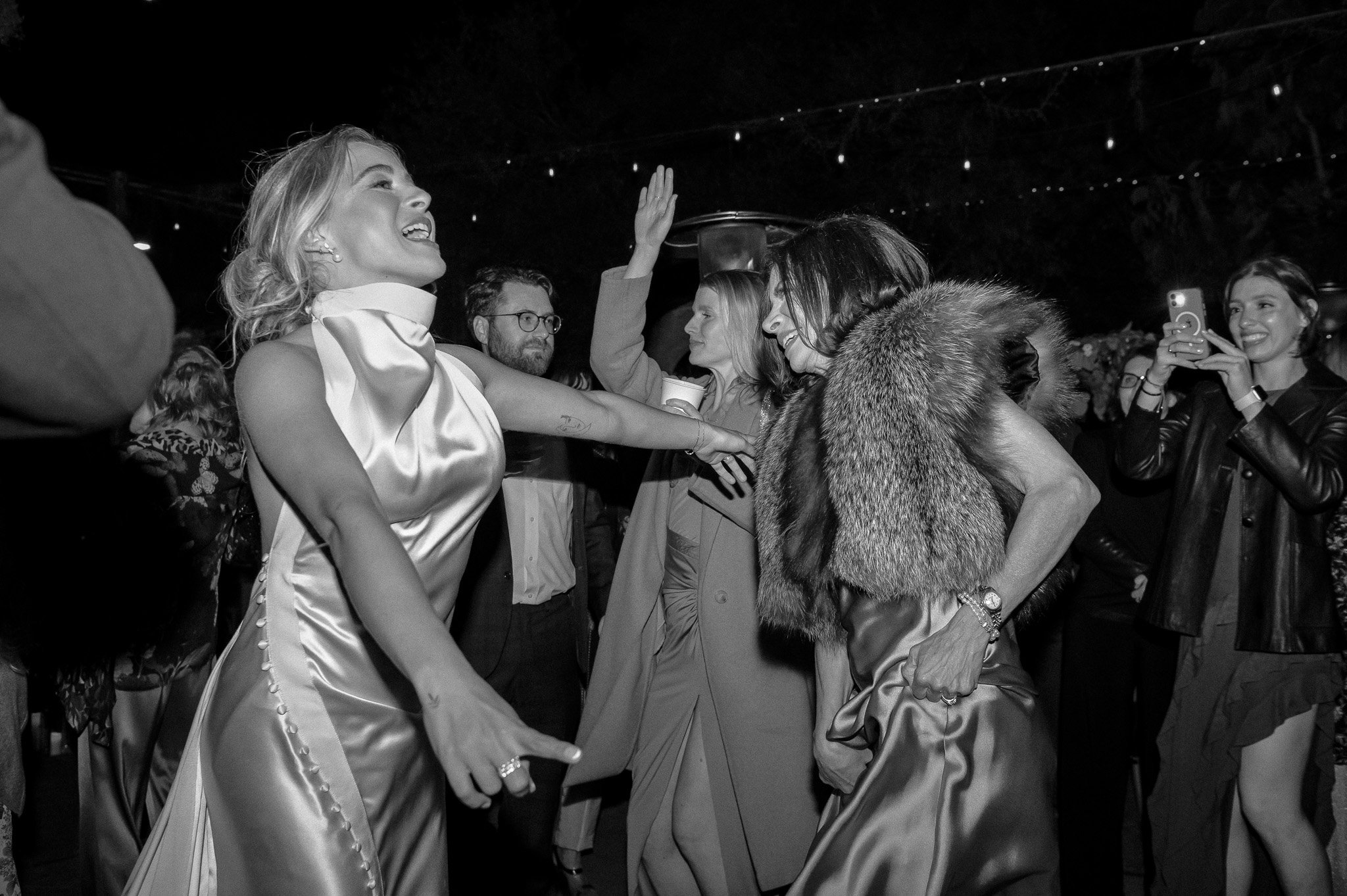 Black and white image of a joyful wedding party dancing at an event, featuring elegantly dressed women in satin and a fur stole, with a crowd enjoying the celebration.