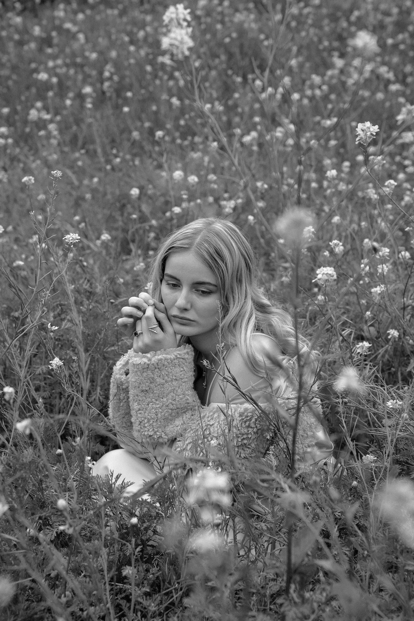 Black and white photo of a woman sitting in a field of flowers, wearing a fluffy jacket, with a contemplative expression.