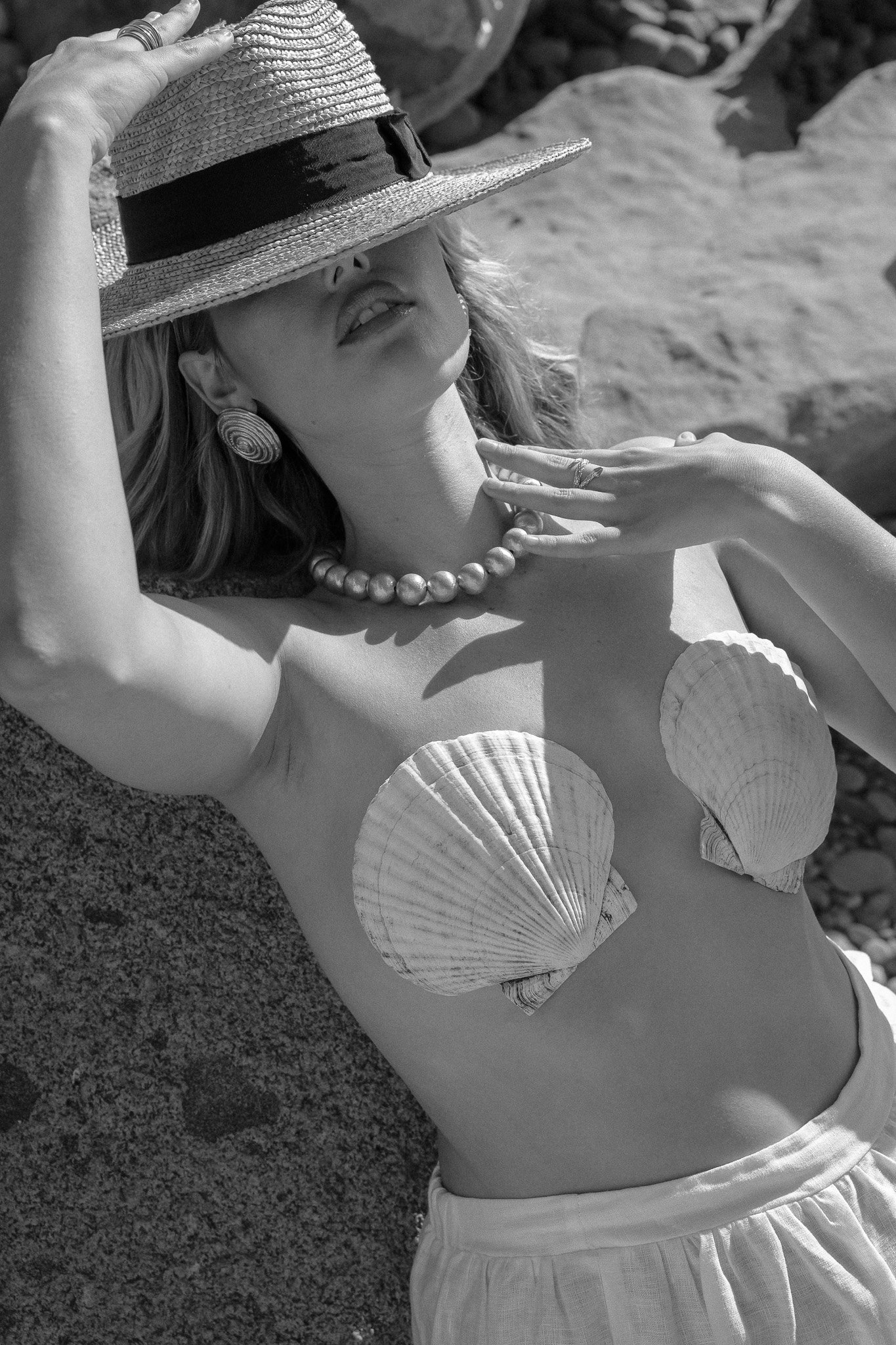 Black and white image of a woman wearing a brimmed hat and a necklace, with seashells as a bikini top, posing in a rocky outdoor setting.