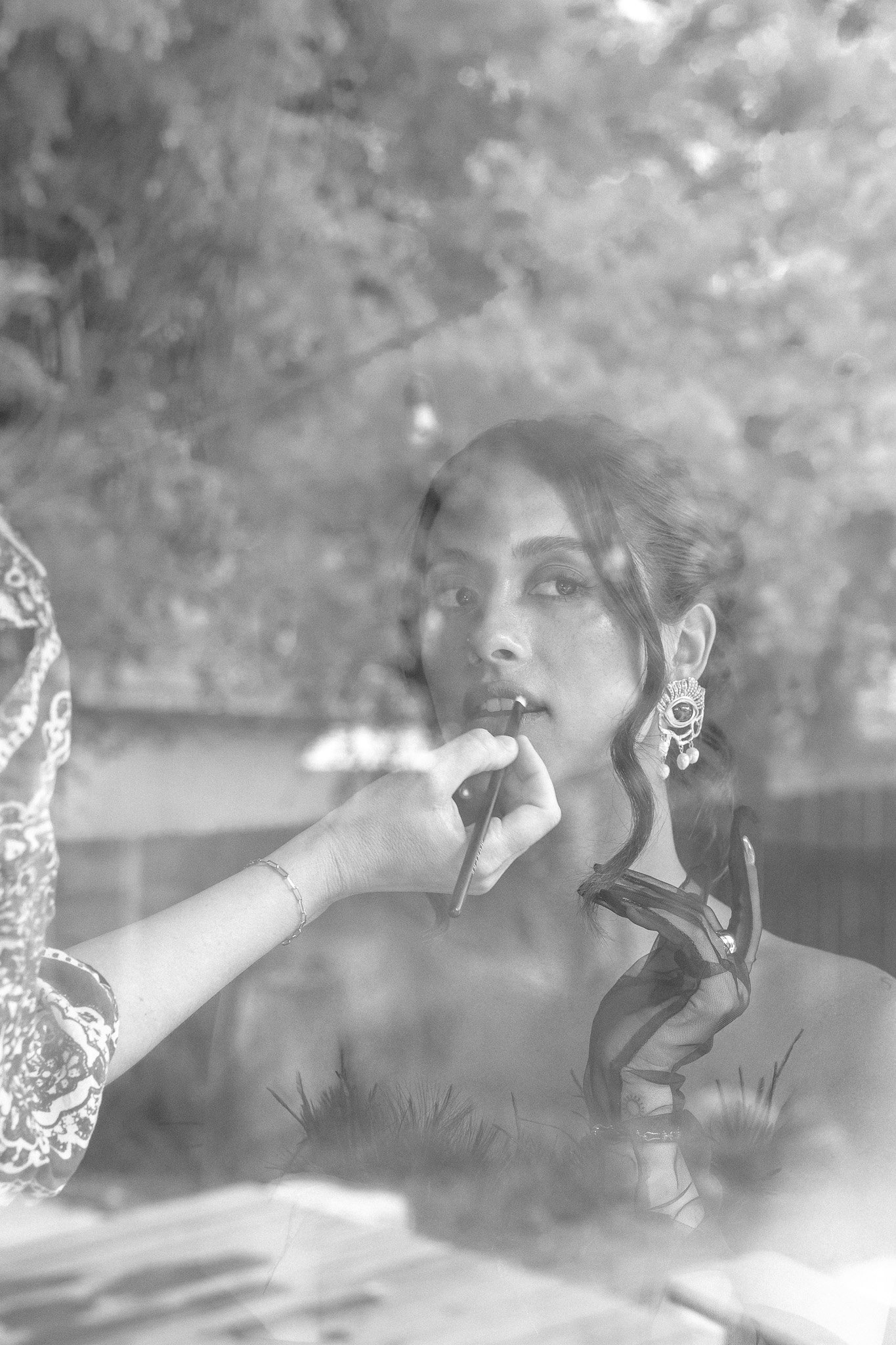 Black and white photo of a woman being applied lipstick, reflection in a window.
