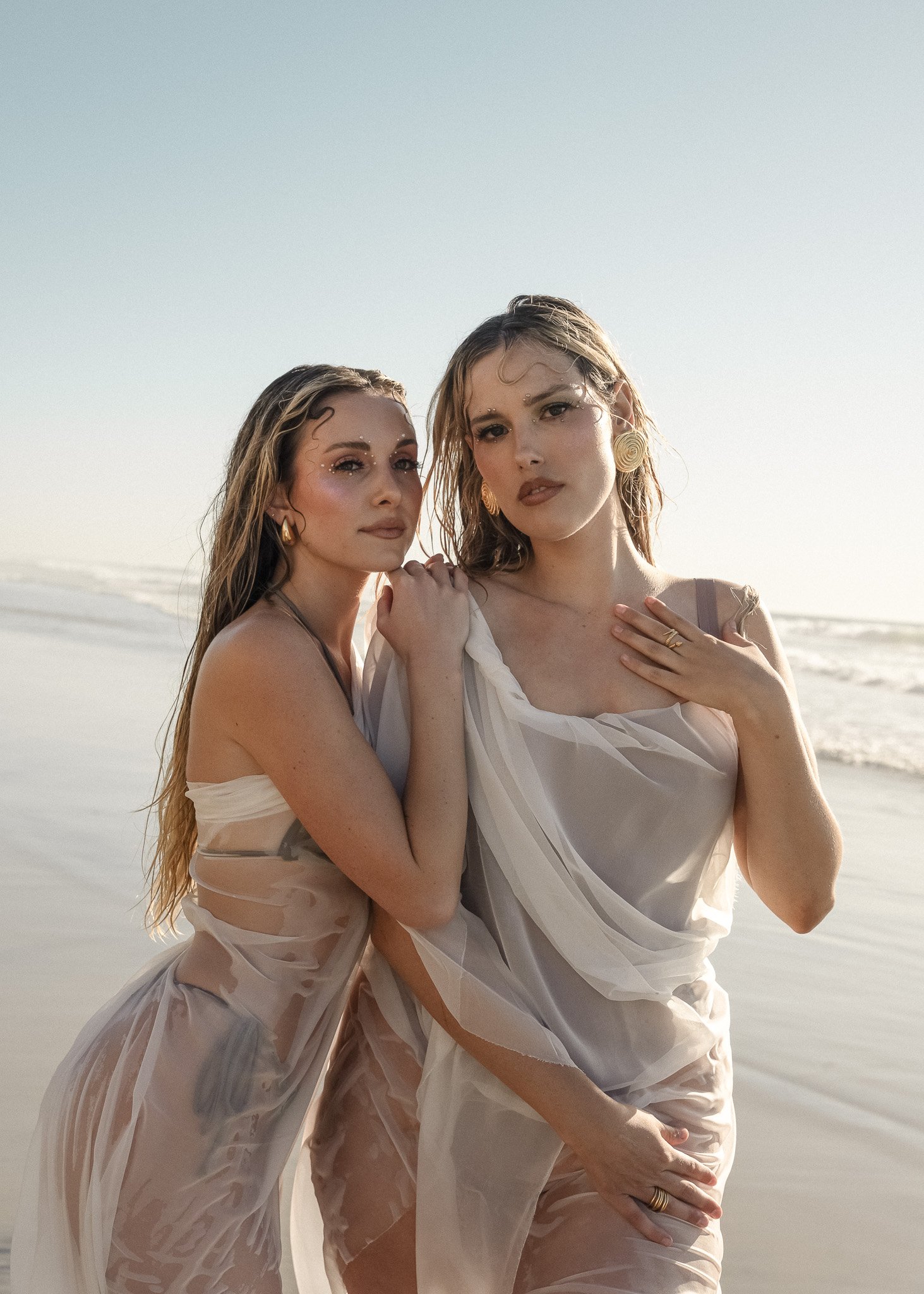 Two women in sheer white dresses, standing on a beach.