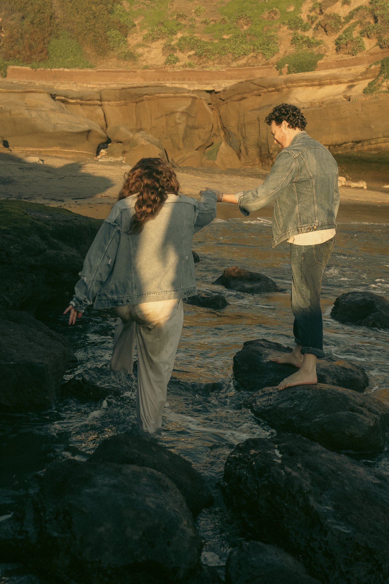 Two people wearing denim jackets walking on rocks near water at the beach.
