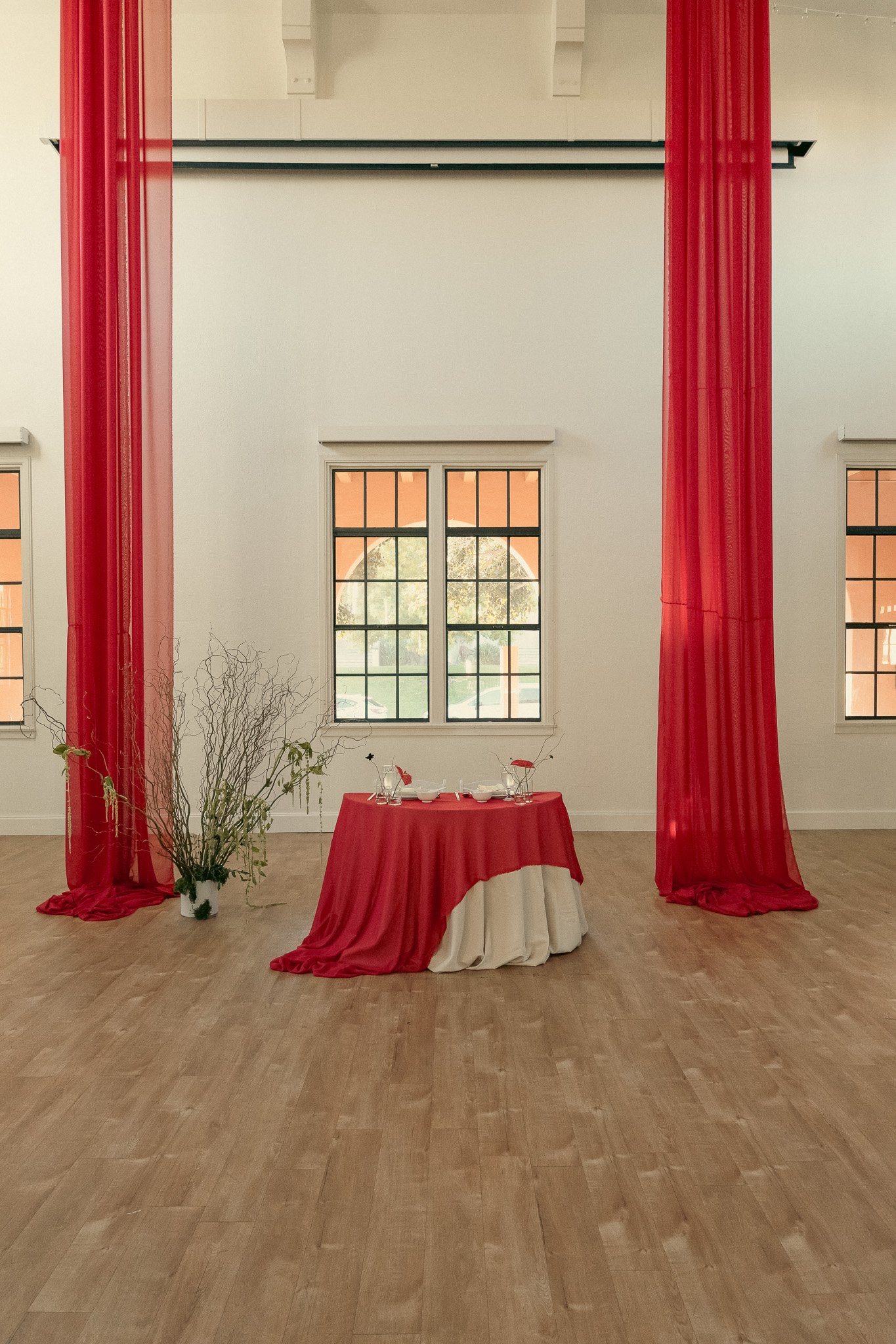 Interior with red drapes, a small table covered with red and white cloth, and sparse plant arrangement in a bright room with wooden floor.