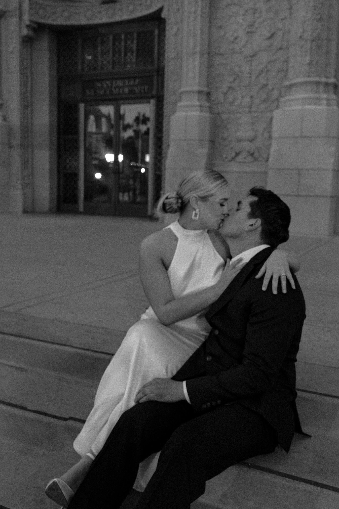 A couple in elegant attire kissing on stone steps in front of a decorative building entrance at night.
