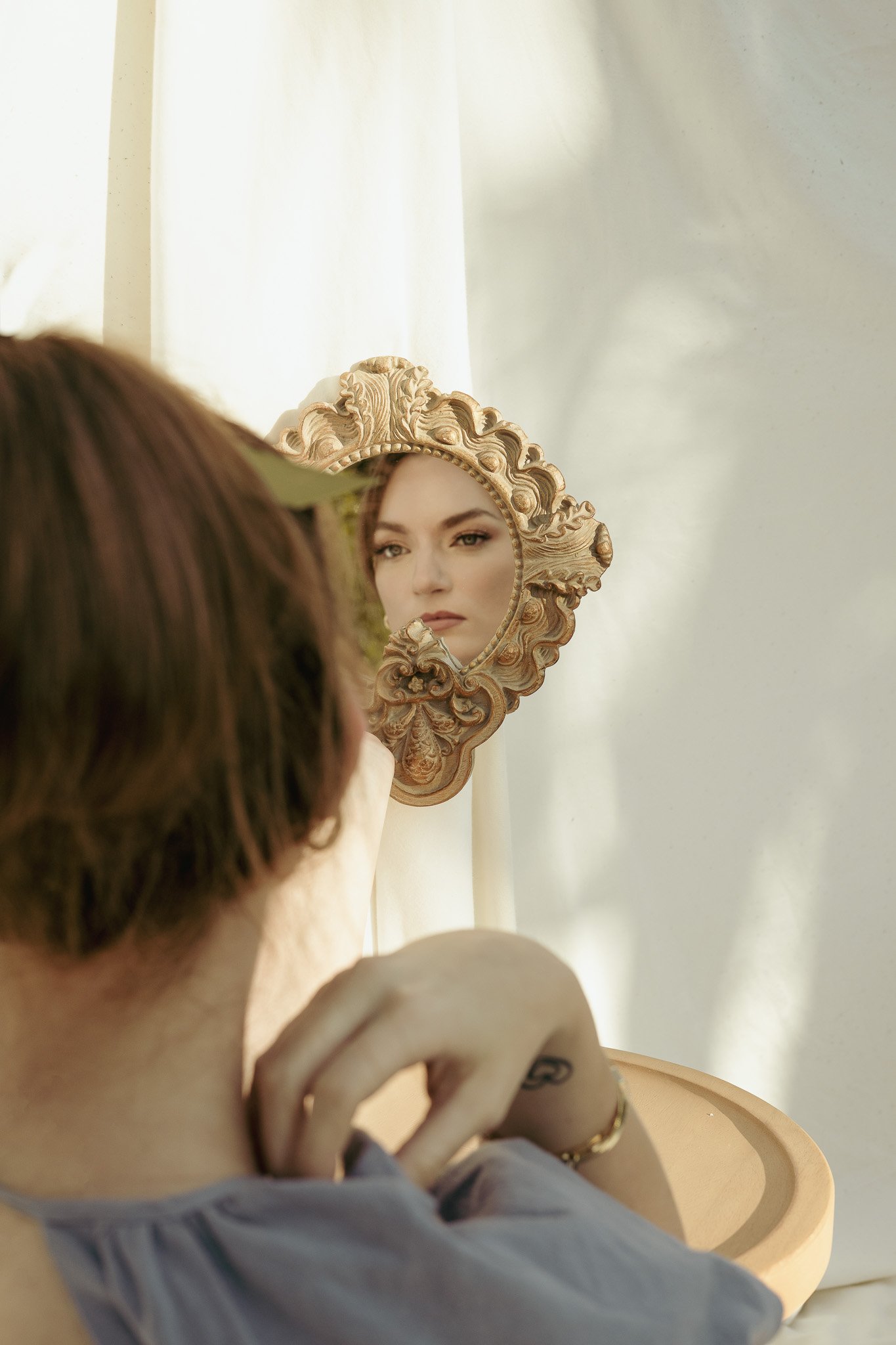 A woman looking into an ornate vintage mirror with a floral design, reflecting her face against a soft, light background.