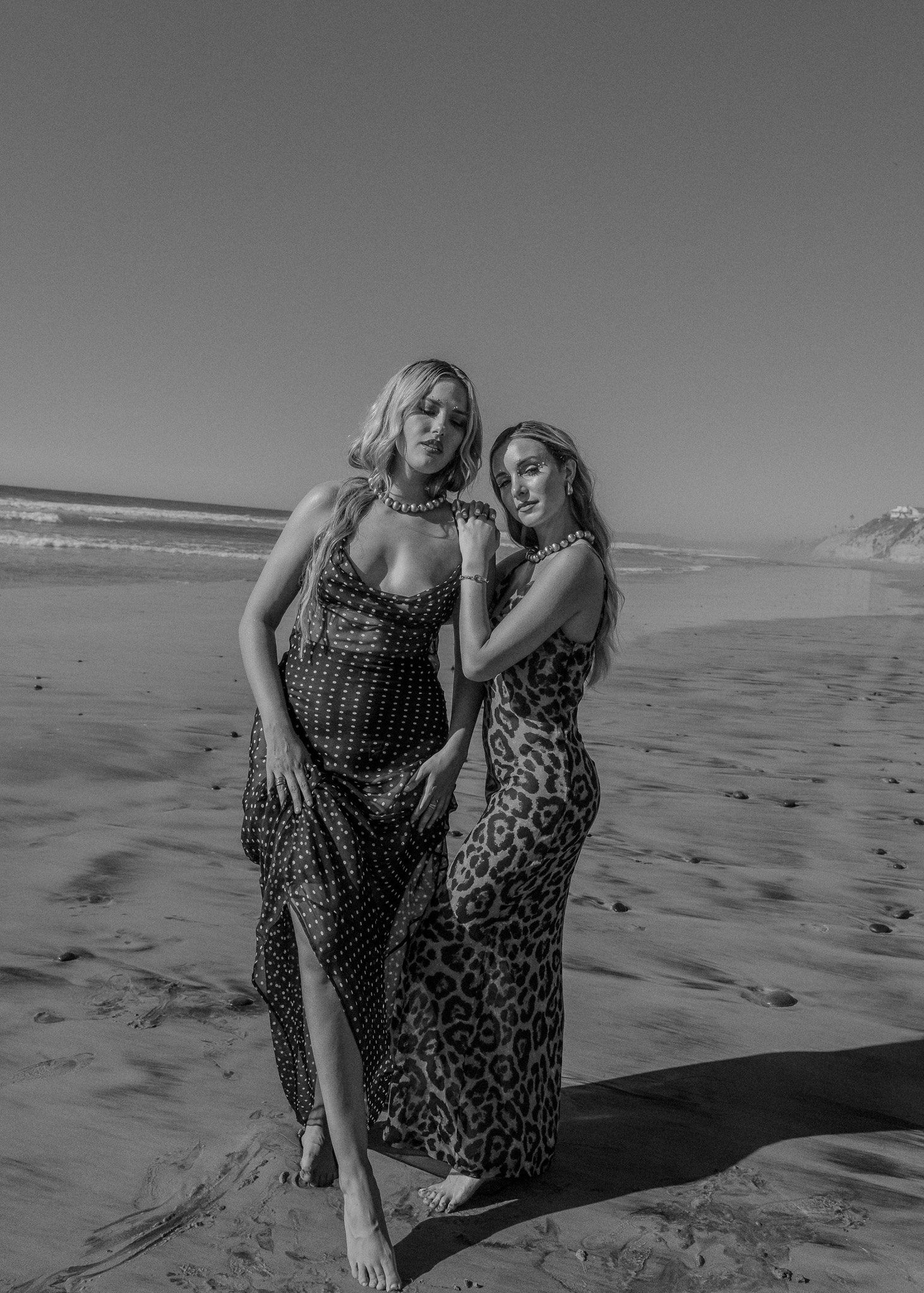 Two women in elegant dresses standing on a beach. One dress is polka-dotted and the other is leopard-print. Both women wear pearl necklaces, and the scene is captured in black and white.