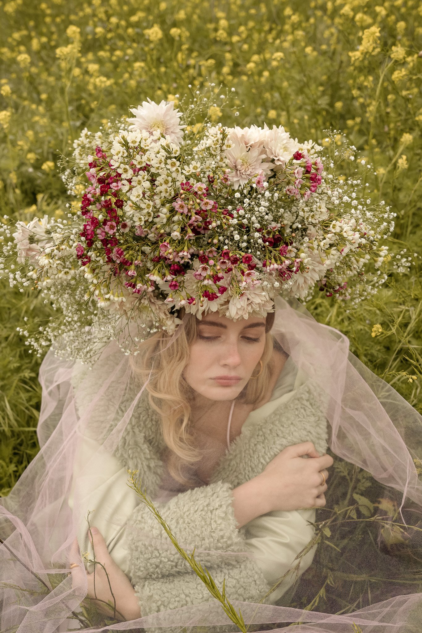 Woman wearing a large floral headpiece and a green coat, sitting in a field of yellow flowers.